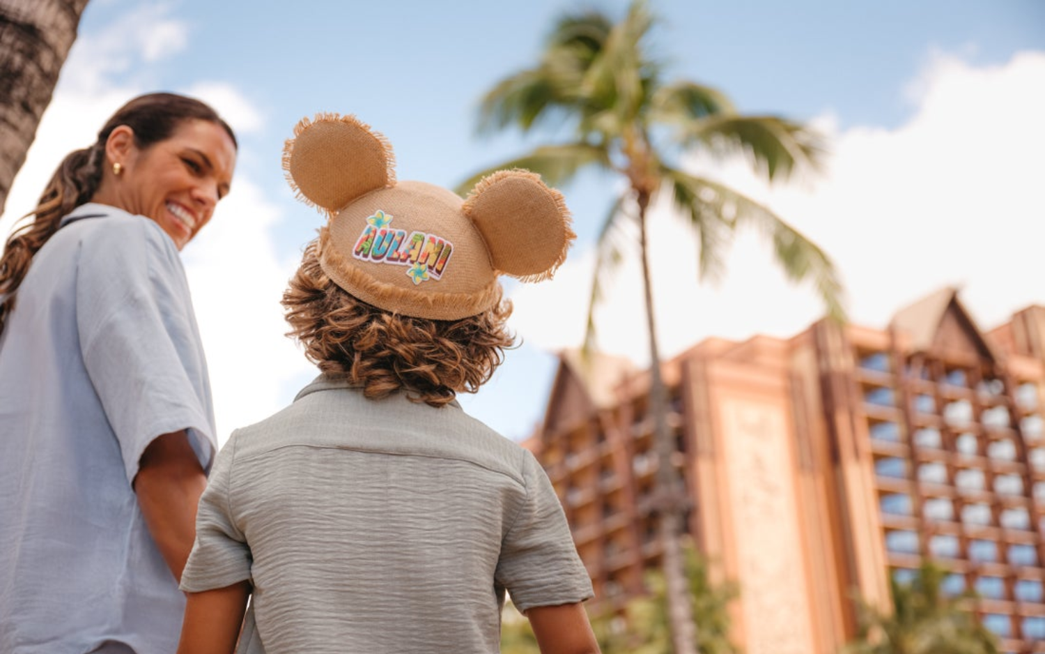 A woman and a child are outdoors. The woman is smiling and looking to the side. The child, seen from behind, wears a hat with ears and colorful lettering that says 'Aulani.' Palm trees and a multi-story building are in the background.