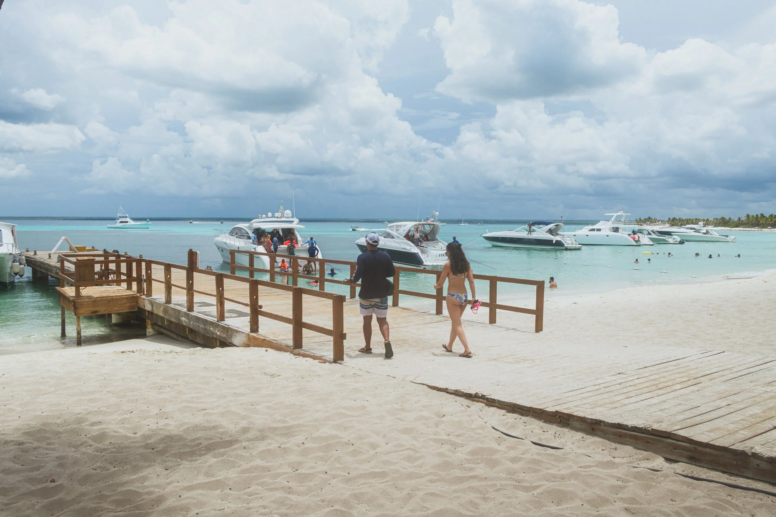 Playa con arena y muelle de madera, personas caminando, barcos yates en el agua turquesa, cielo nublado.