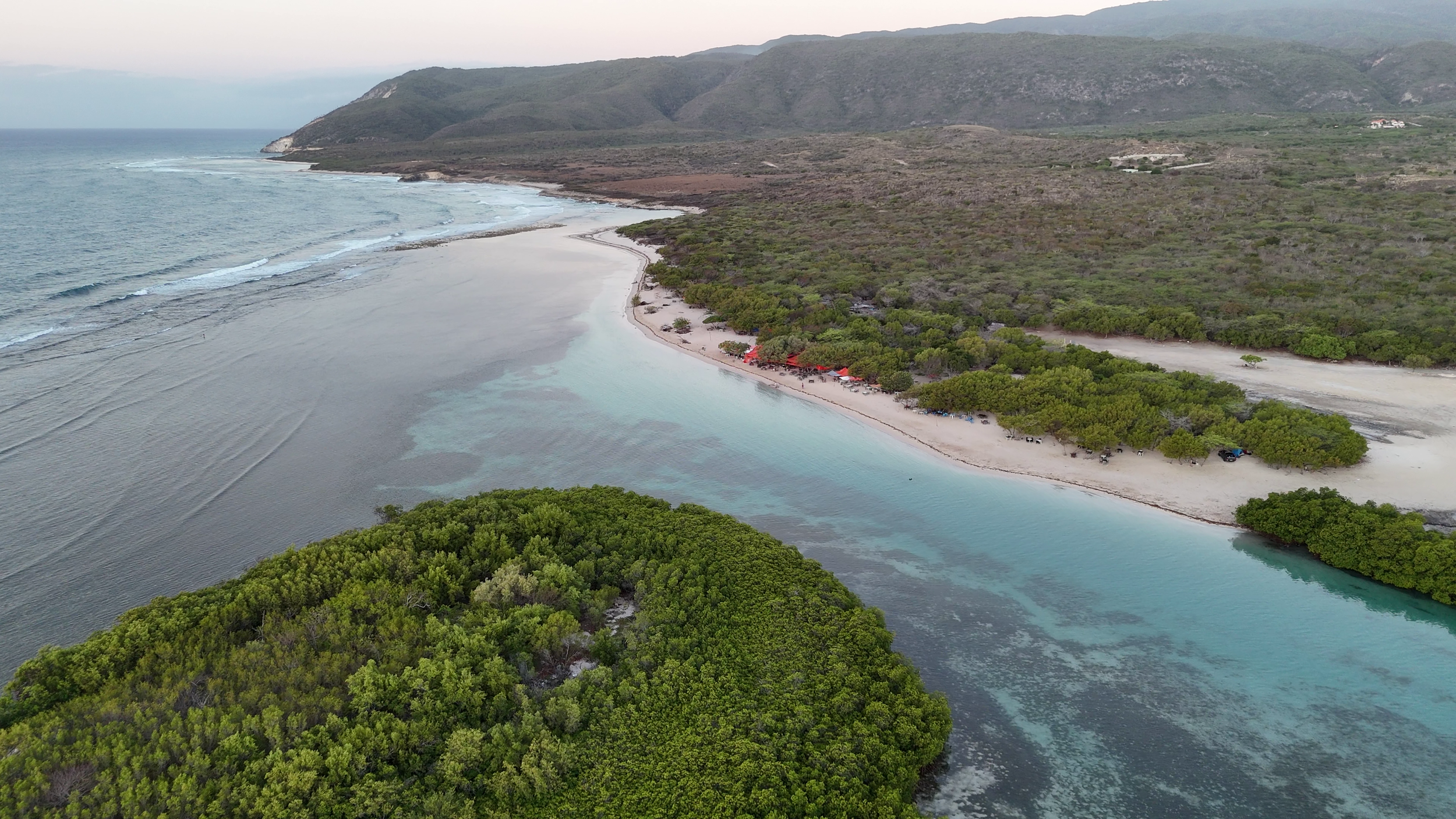 Vista aérea de una playa con agua turquesa y áreas de manglares, rodeada de vegetación y montañas al fondo. El mar se extiende hacia el horizonte.