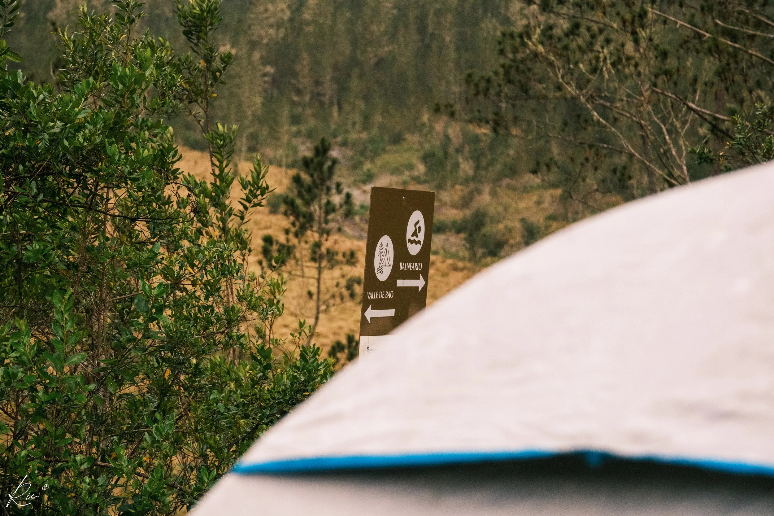 Paisaje natural con un cartel que indica direcciones hacia "Valle de Bao" y "Balneario" rodeado de vegetación y árboles.