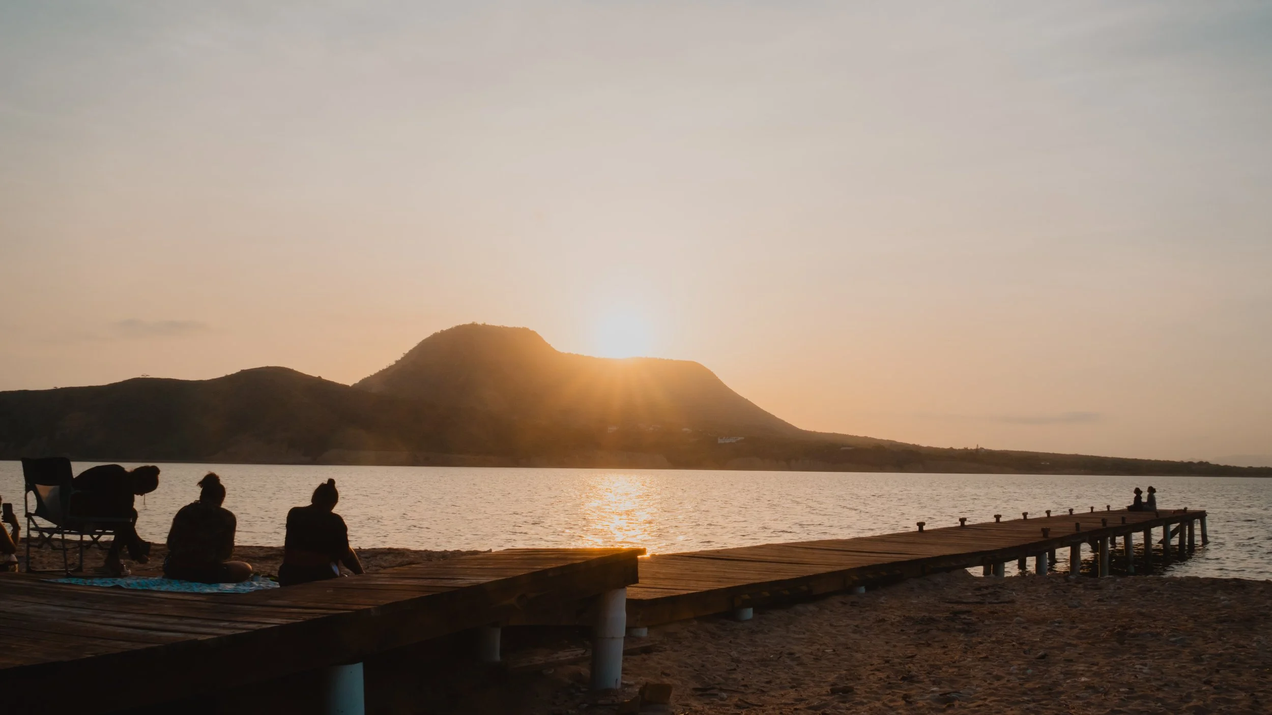 Personas viendo la puesta de sol desde un muelle, con un lago y montañas al fondo.