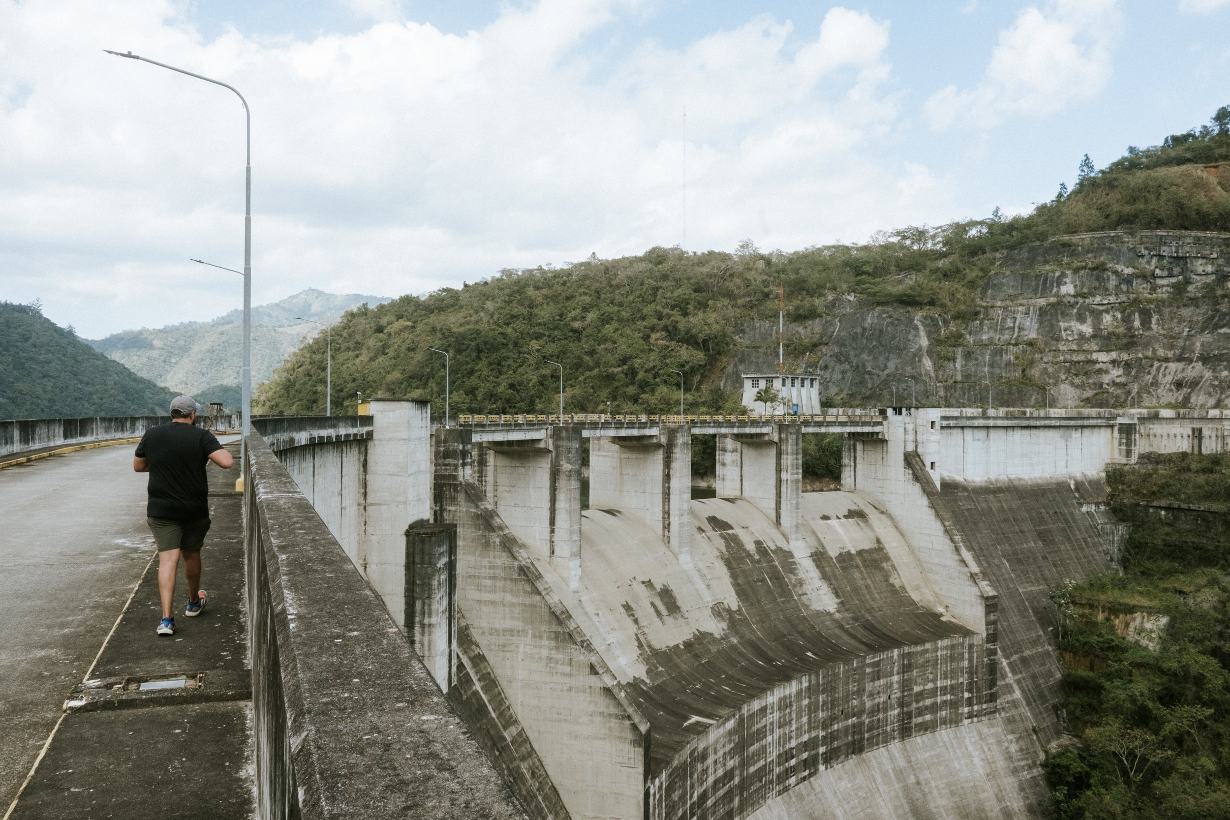 Persona caminando sobre una presa de concreto con montañas y vegetación en el fondo.