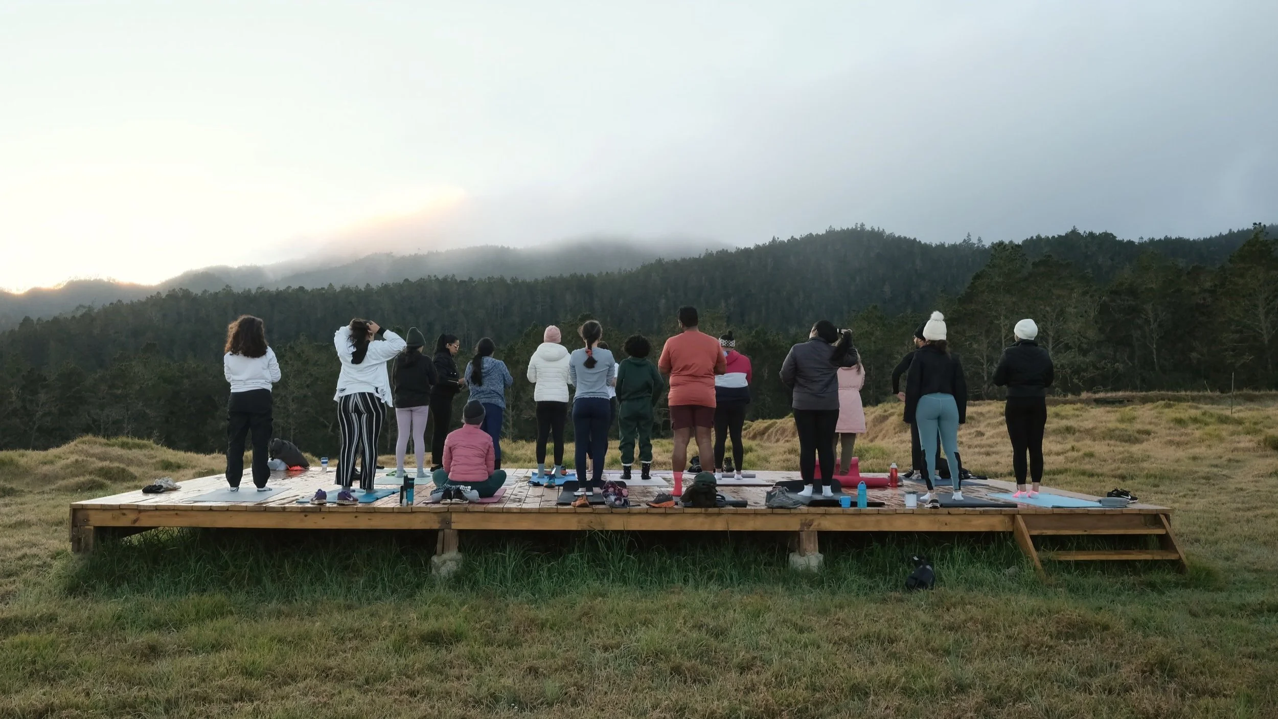 Grupo de personas haciendo yoga al aire libre sobre una plataforma de madera en un entorno natural con montañas y árboles de fondo.
