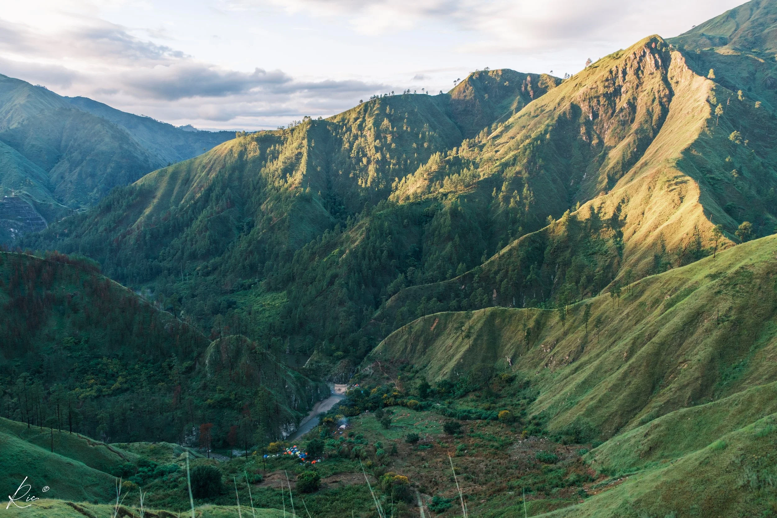 Vista panorámica de un paisaje montañoso verde iluminado por el sol, con sombras alargadas. Se observa un pequeño valle con vegetación densa y una ligera pista serpenteando entre las montañas.