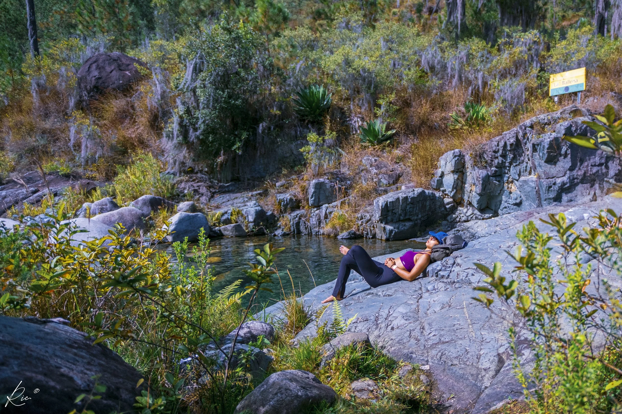 Persona descansando sobre una roca cerca de un río en un entorno natural, rodeada de vegetación.