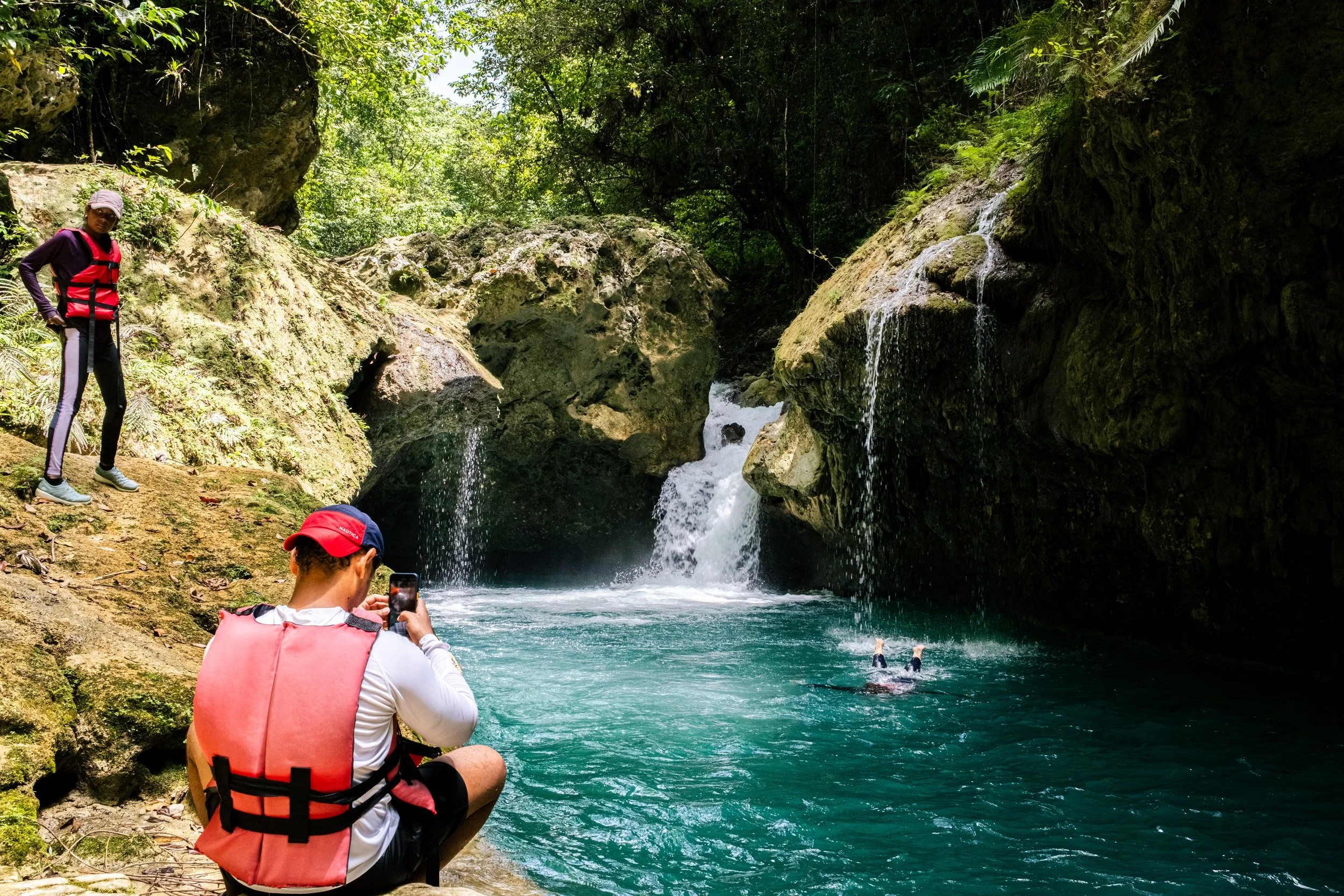 Personas en chaleco salvavidas en un entorno natural con cascadas y un estanque de agua azul, rodeados de vegetación y rocas.