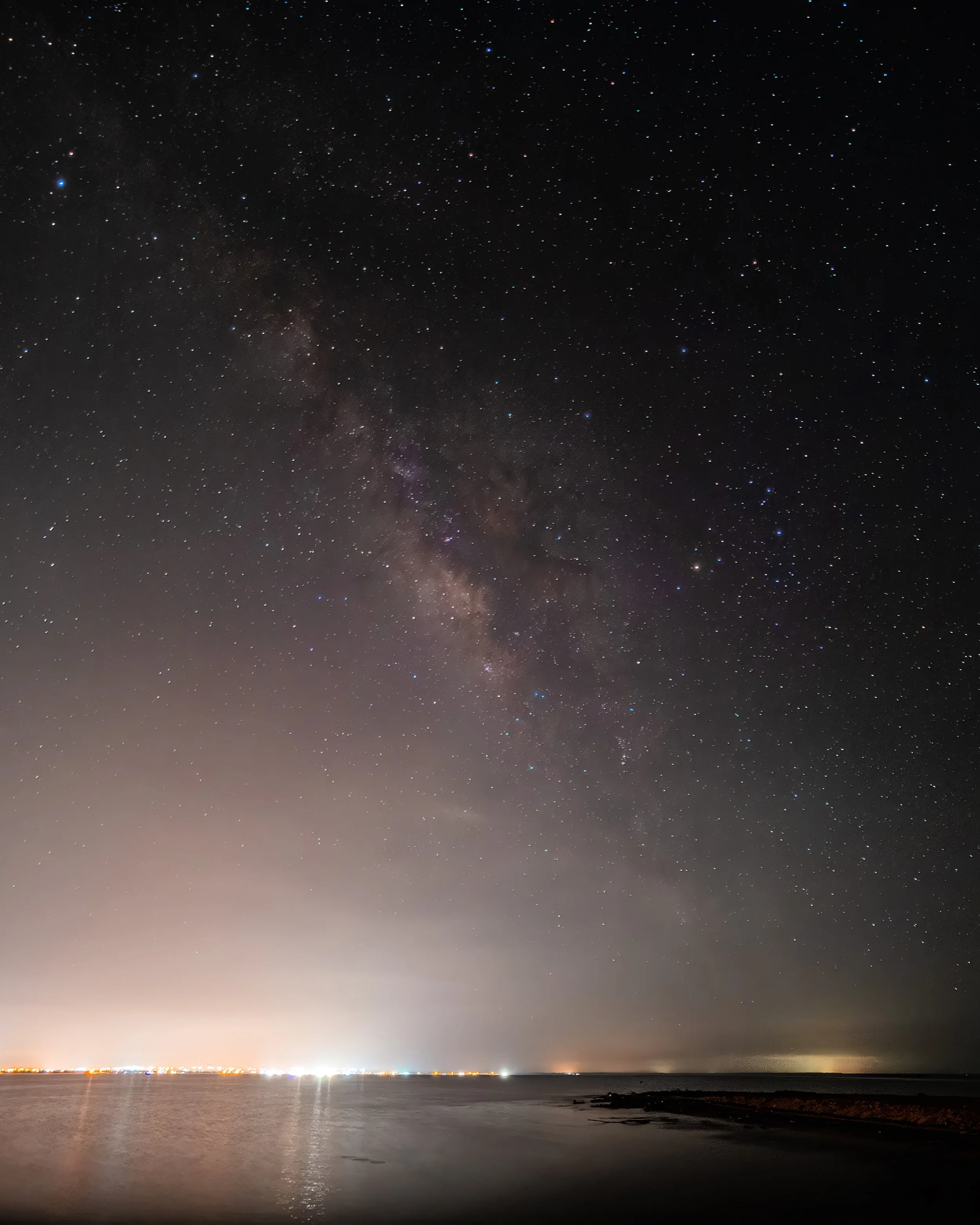 Cielo estrellado con la Vía Láctea sobre un paisaje costero nocturno, luces de la ciudad visibles en el horizonte.