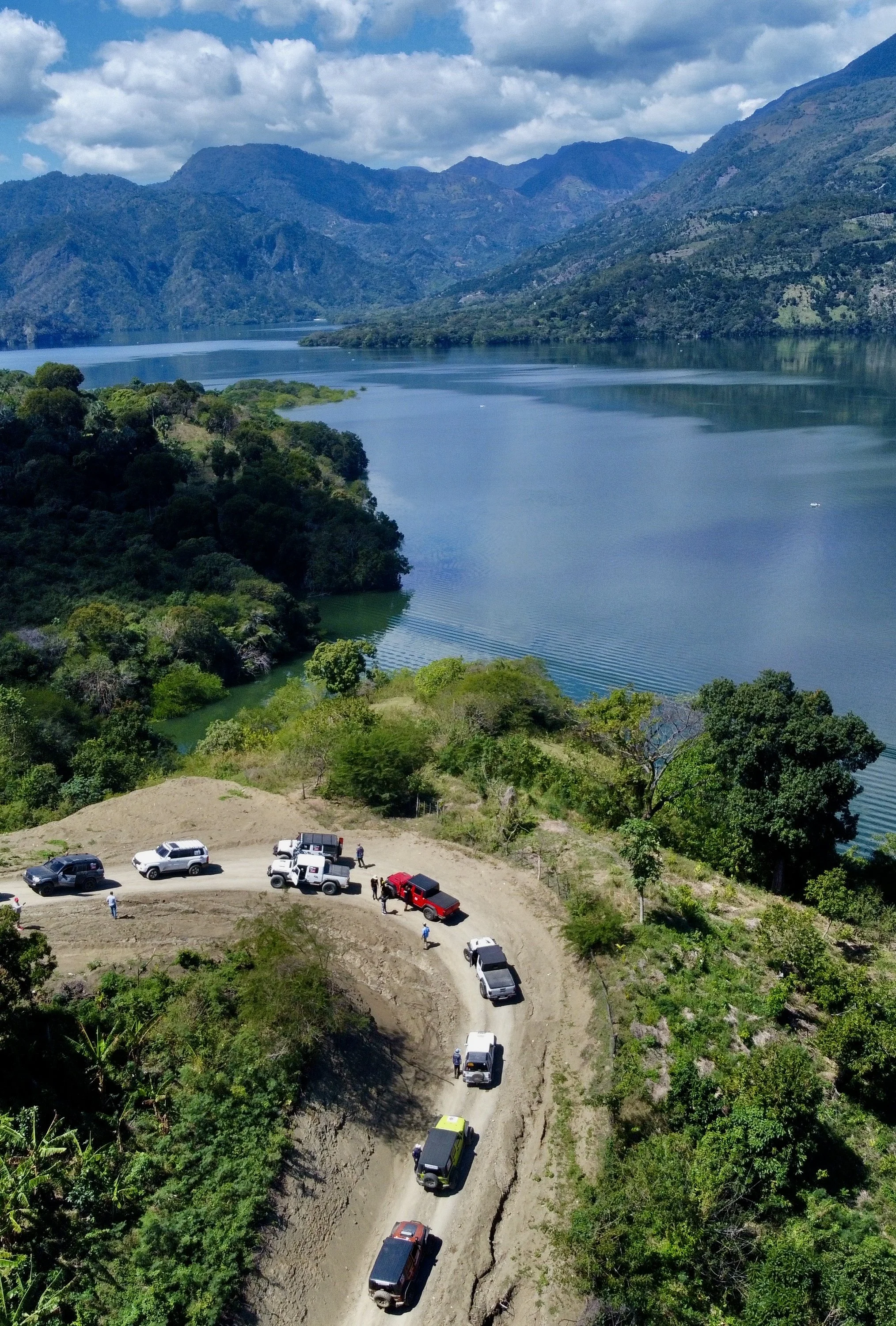 Carretera sinuosa con varios vehículos y personas en un entorno montañoso junto a un lago.