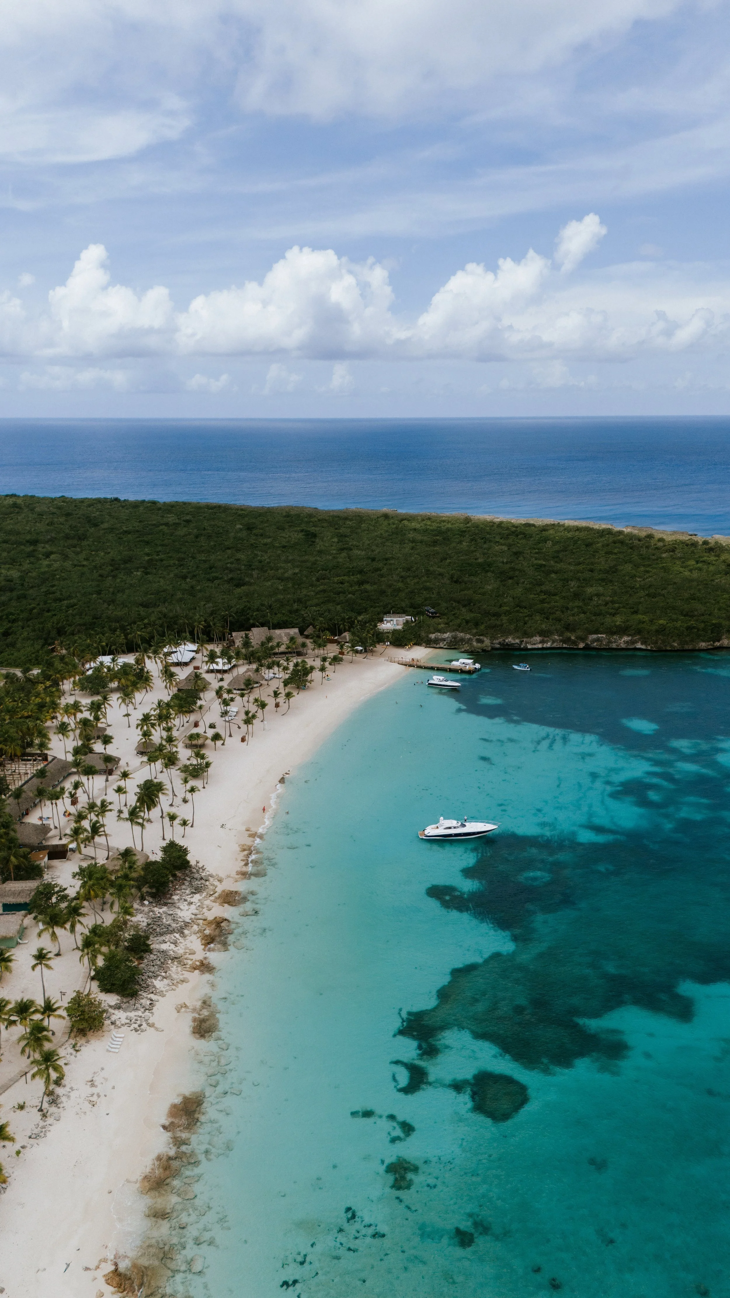 Playa tropical con arena blanca, palmeras y agua azul cristalina, vista aérea. Hay barcos en el agua junto a la orilla. Vegetación y bosque al fondo.