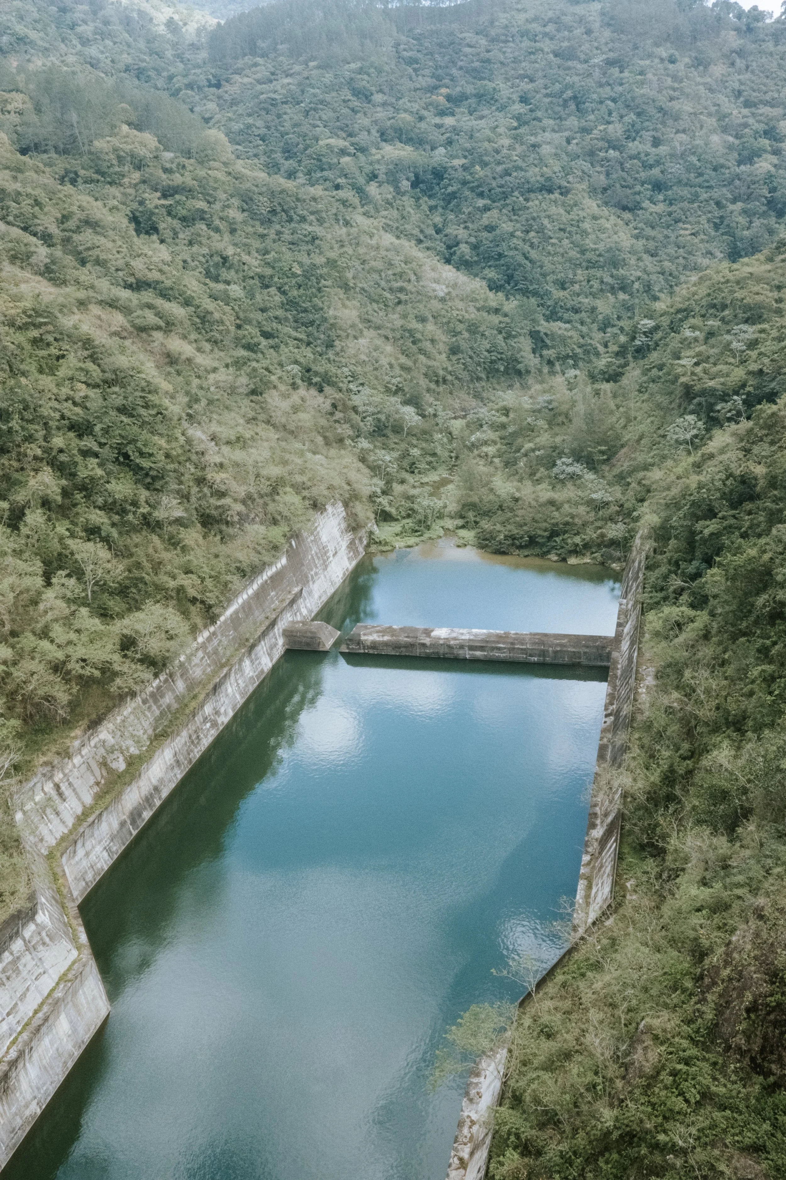 Vista aérea de un embalse entre colinas verdes, con agua azul clara y paredes de concreto, rodeado de vegetación abundante.