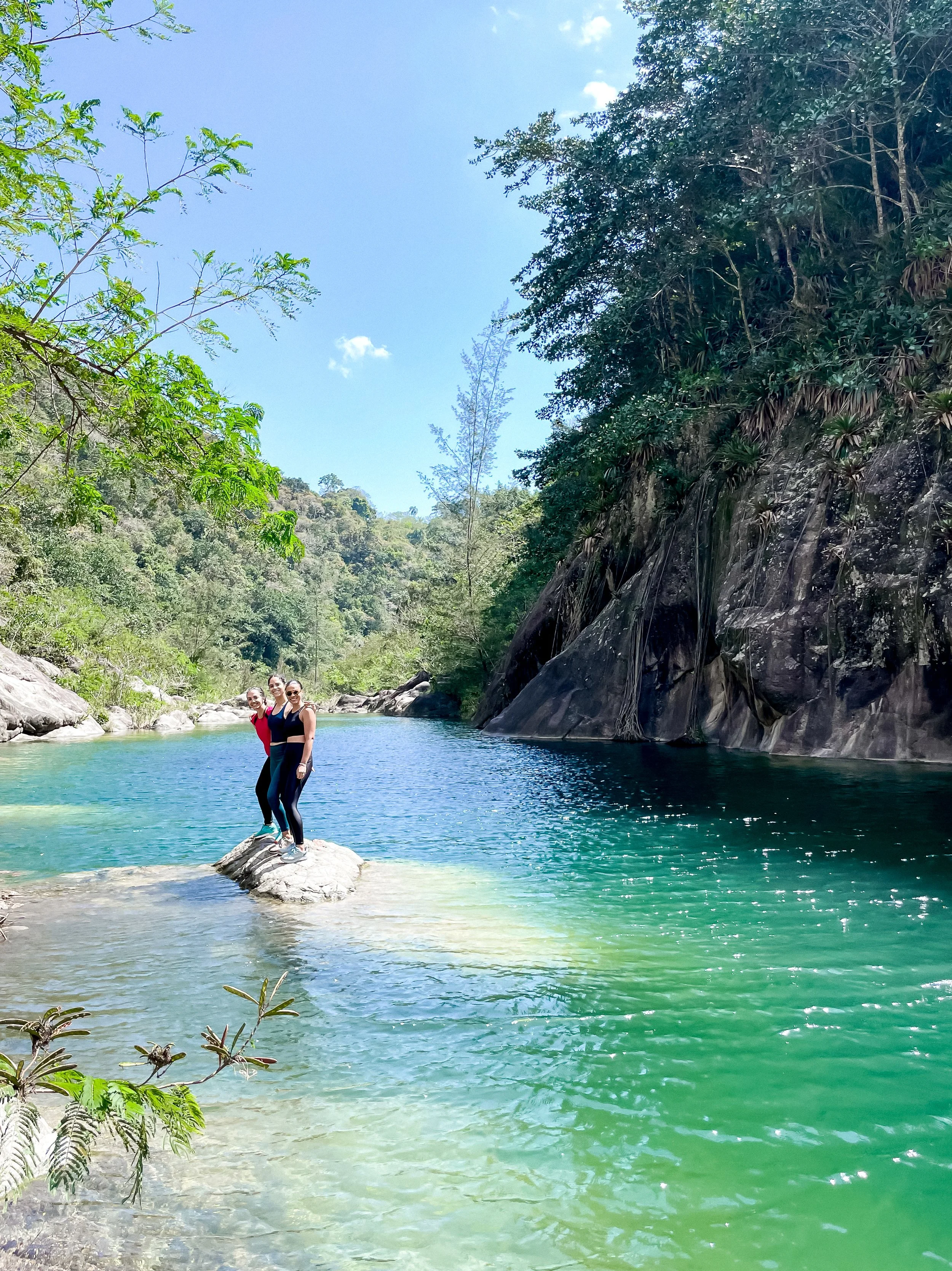 Dos personas posan sobre una roca en medio de un río de aguas cristalinas rodeado de vegetación y montañas, bajo un cielo despejado.