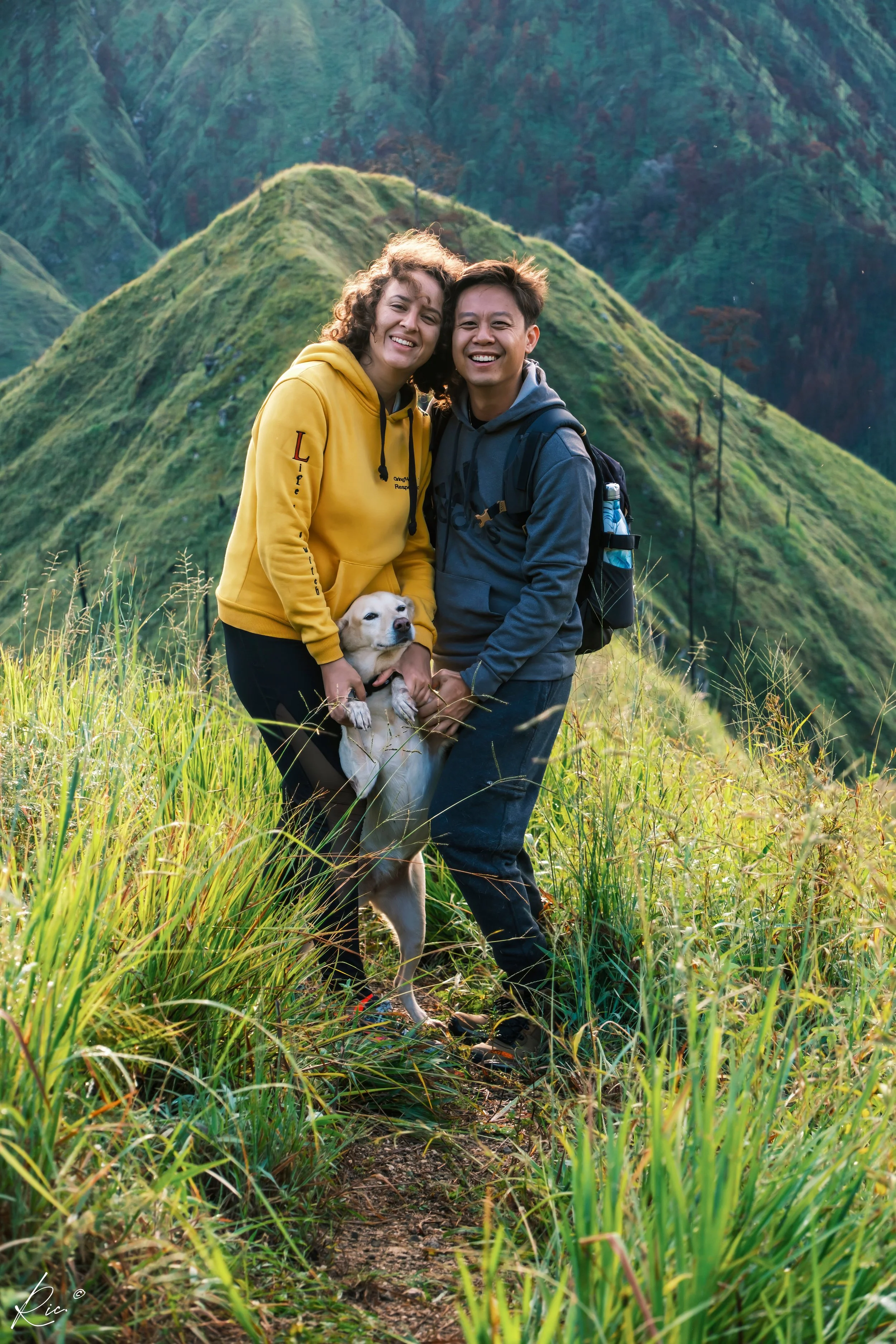 Dos personas sonriendo mientras sostienen a un perro en medio de un paisaje montañoso verde.