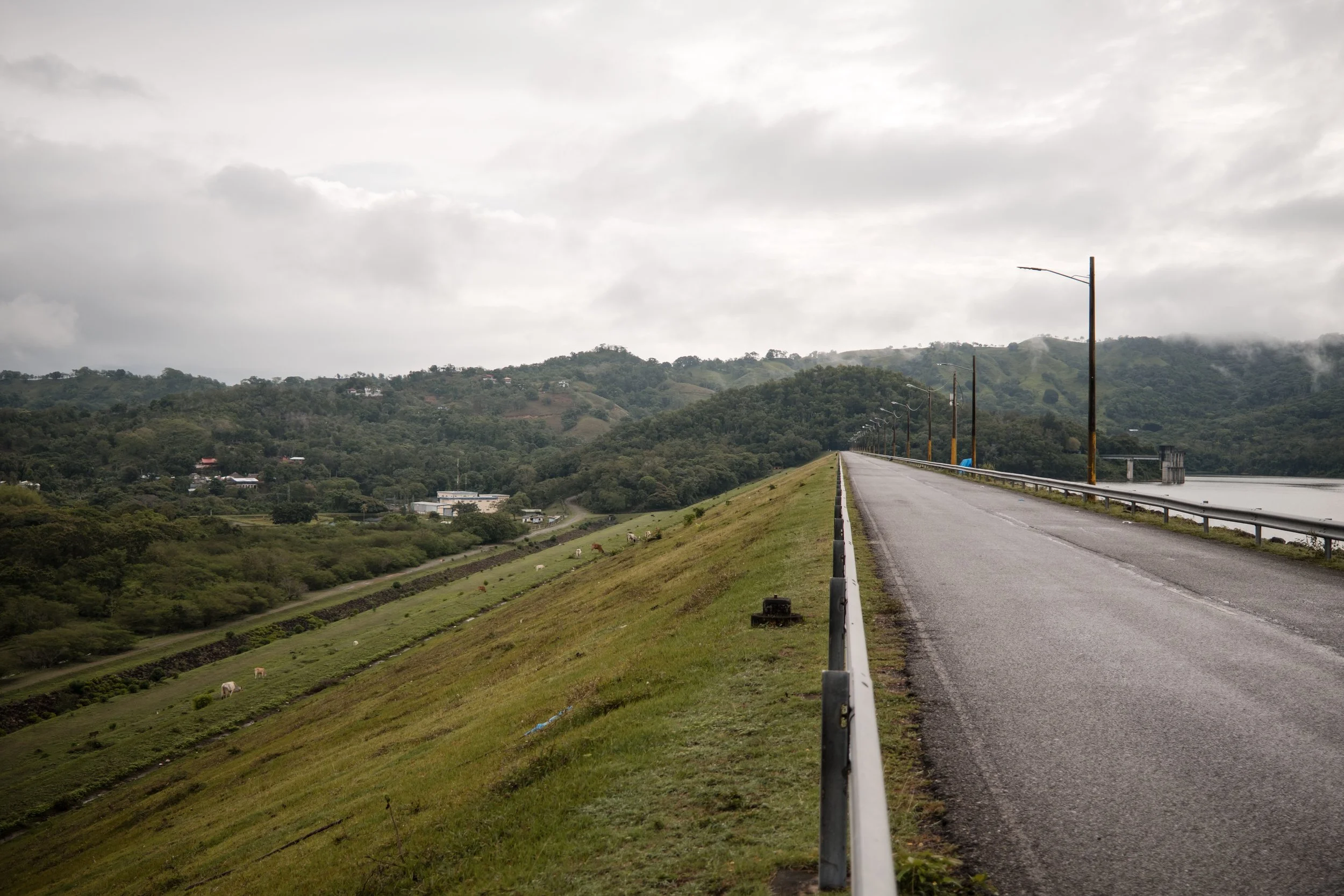Carretera junto a un embalse, con colinas verdes y nubes en el cielo.