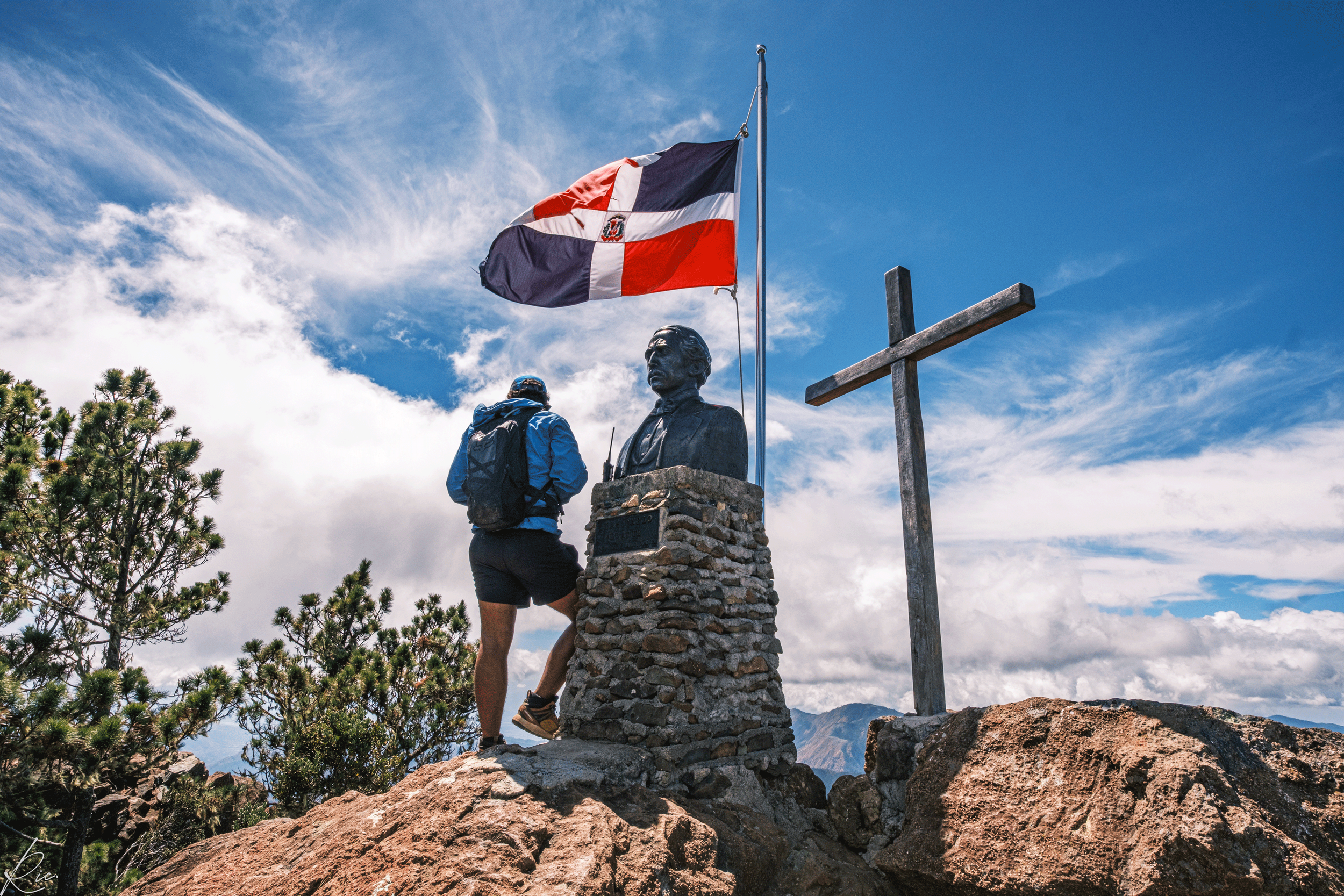Persona en montaña junto a busto, bandera dominicana y cruz.