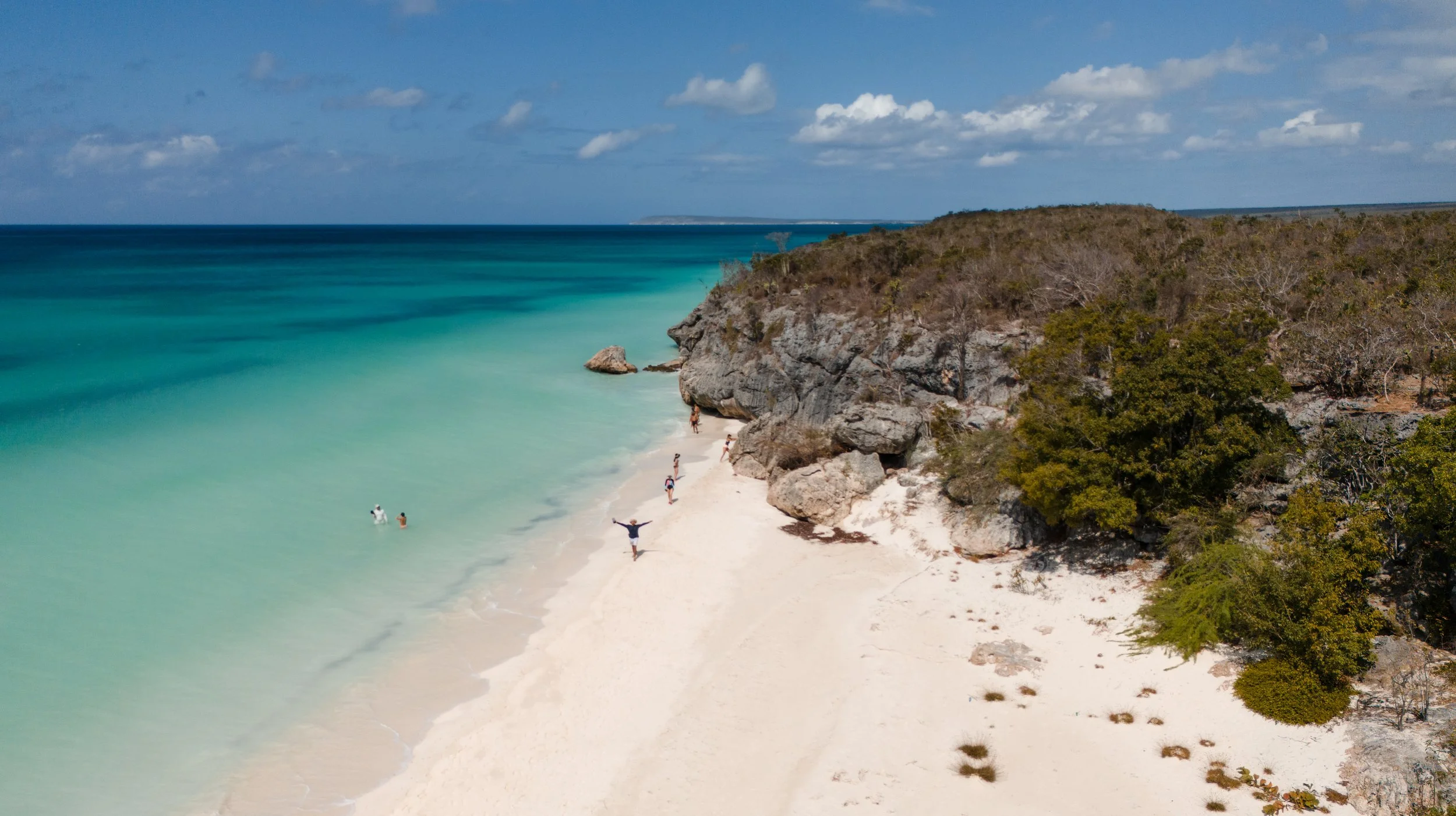 Playa con agua turquesa, arena blanca y acantilados con vegetación. Personas caminando y nadando en el mar bajo un cielo despejado.