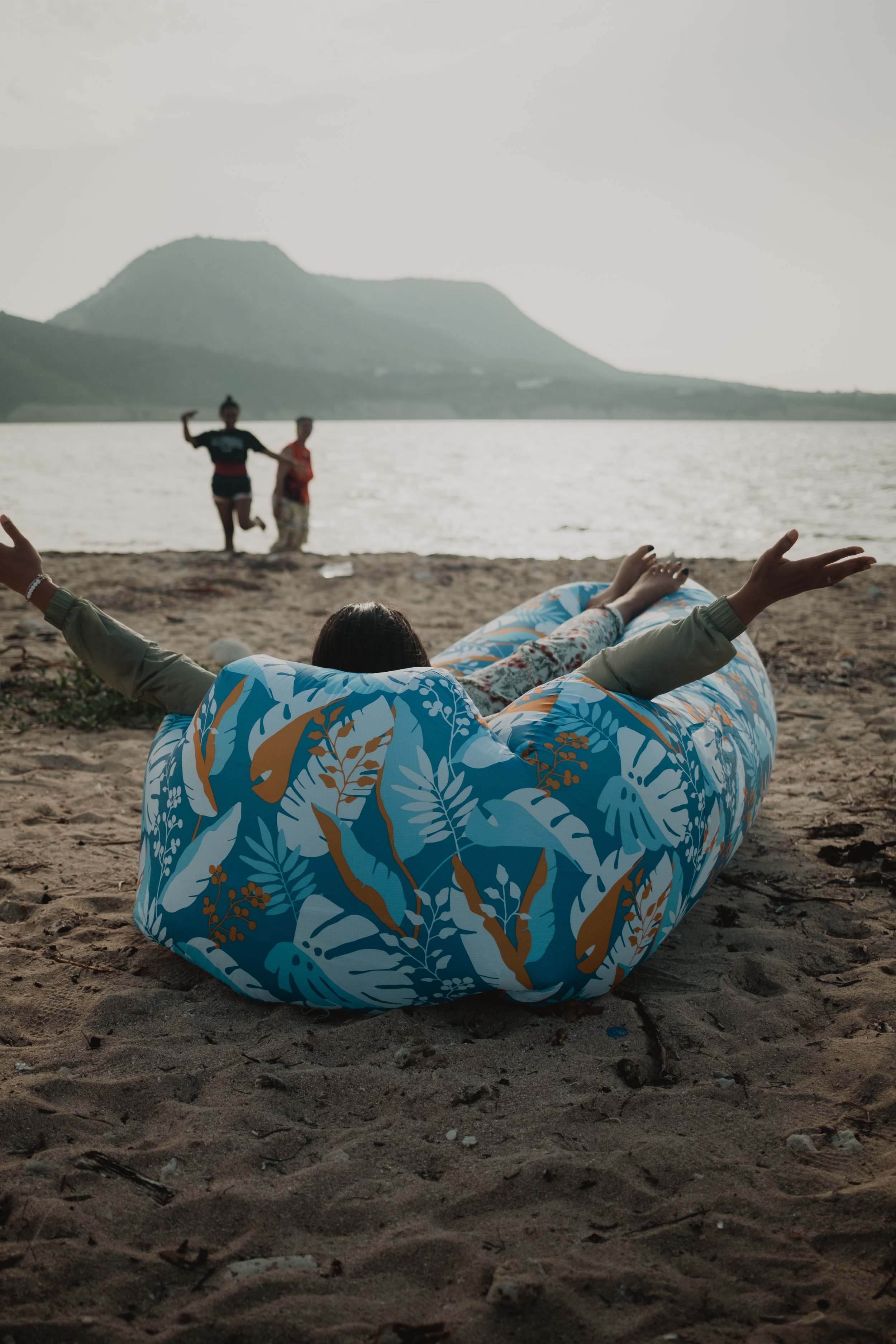 Persona relajándose en una playa sobre un colchón inflable con estampado colorido, con montañas y mar de fondo. Dos personas caminando en la distancia.
