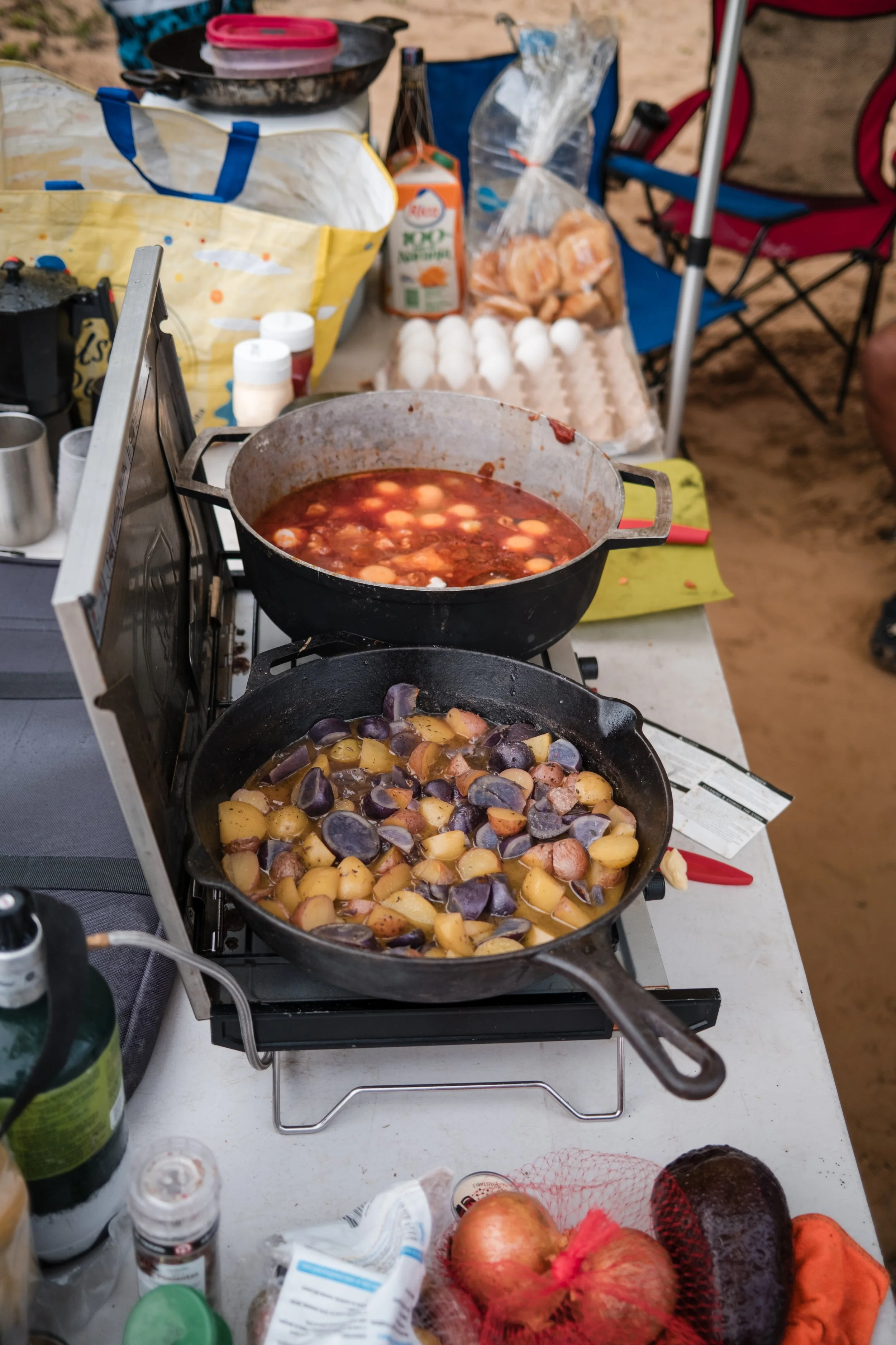 Preparación de comida al aire libre con dos cacerolas: una con papas de colores y otra con un guiso rojo. Huevos, pan y otros ingredientes en la mesa.