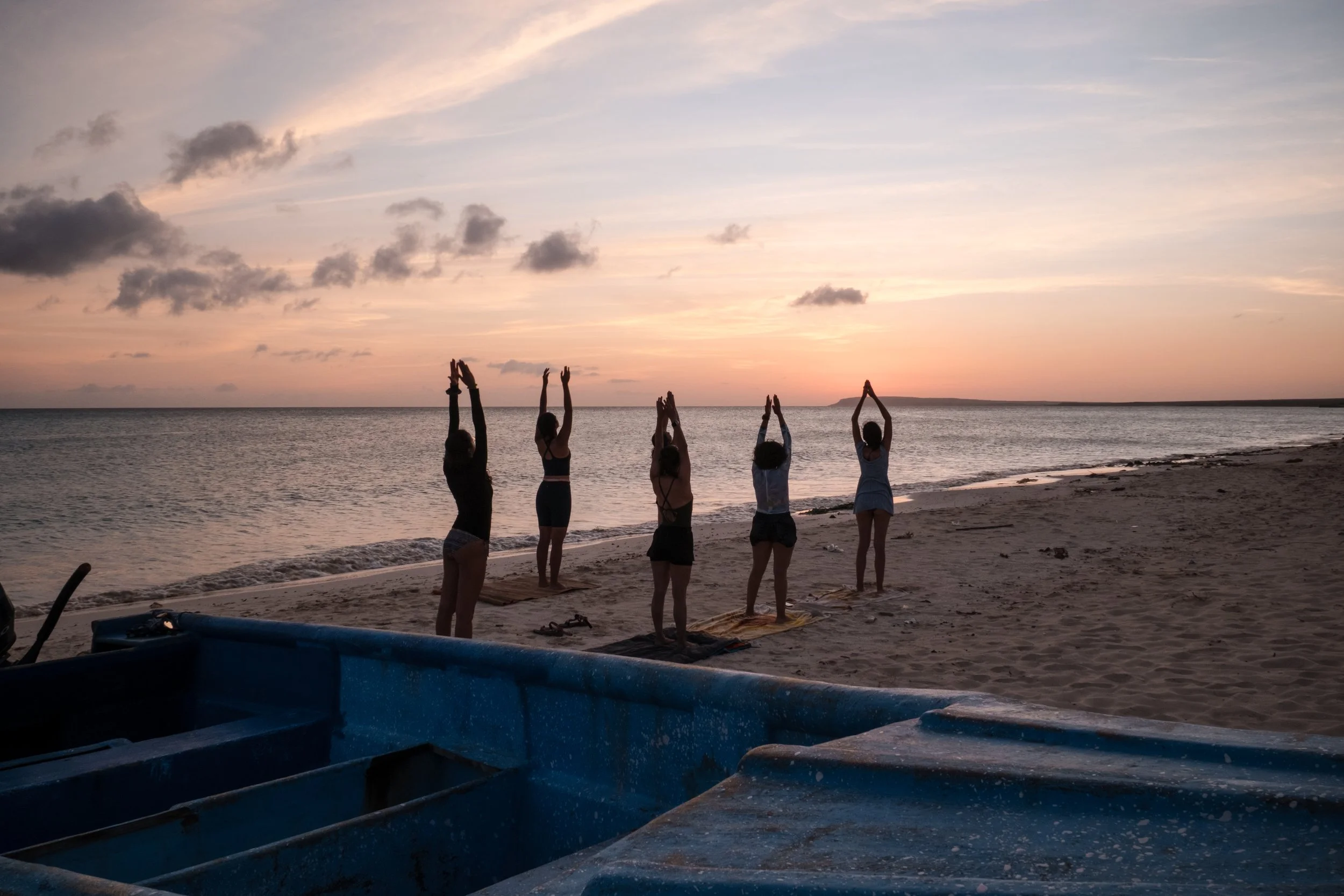 Grupo de personas haciendo yoga al atardecer en la playa, con un bote azul en primer plano.