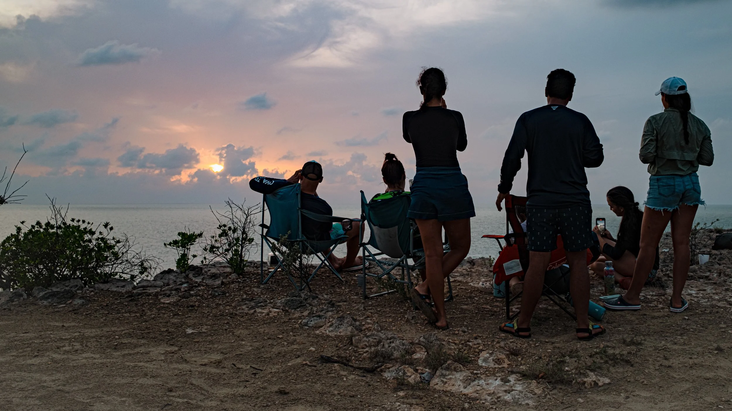 Grupo de personas mirando el atardecer en la playa, sentados y de pie, con cielo nublado y mar al fondo.