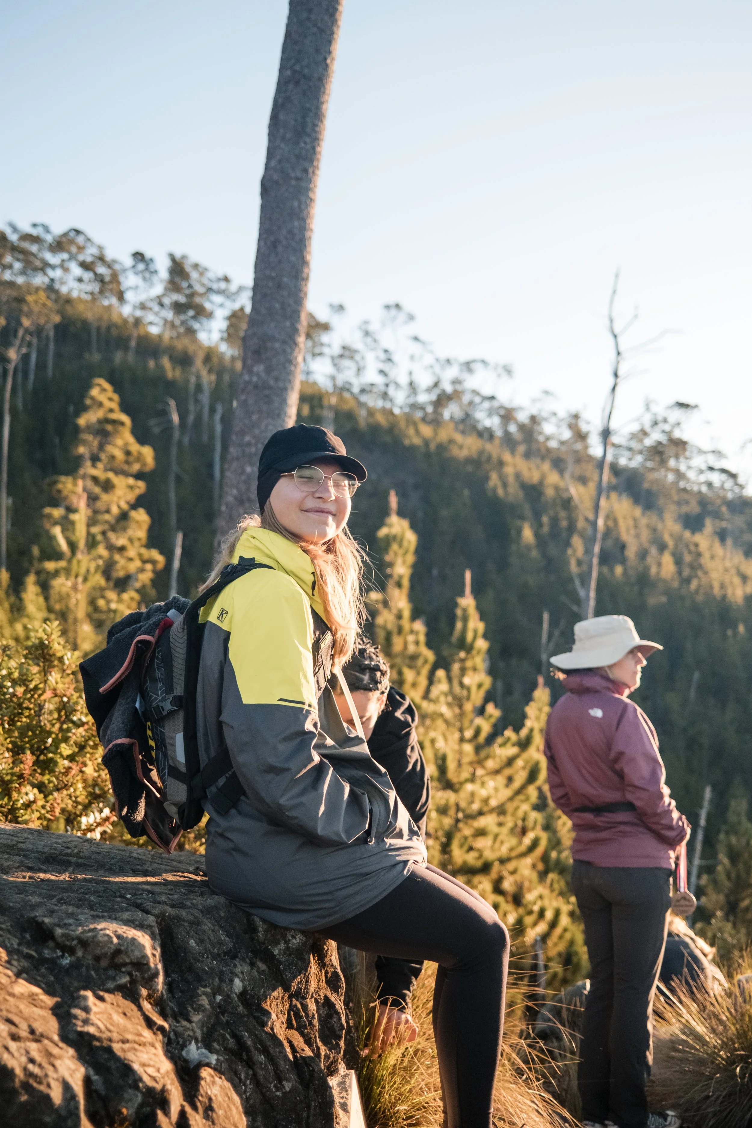 Grupo de excursionistas en una montaña, con árboles de fondo y luz de sol. Una persona sentada lleva chaqueta amarilla y mochila.
