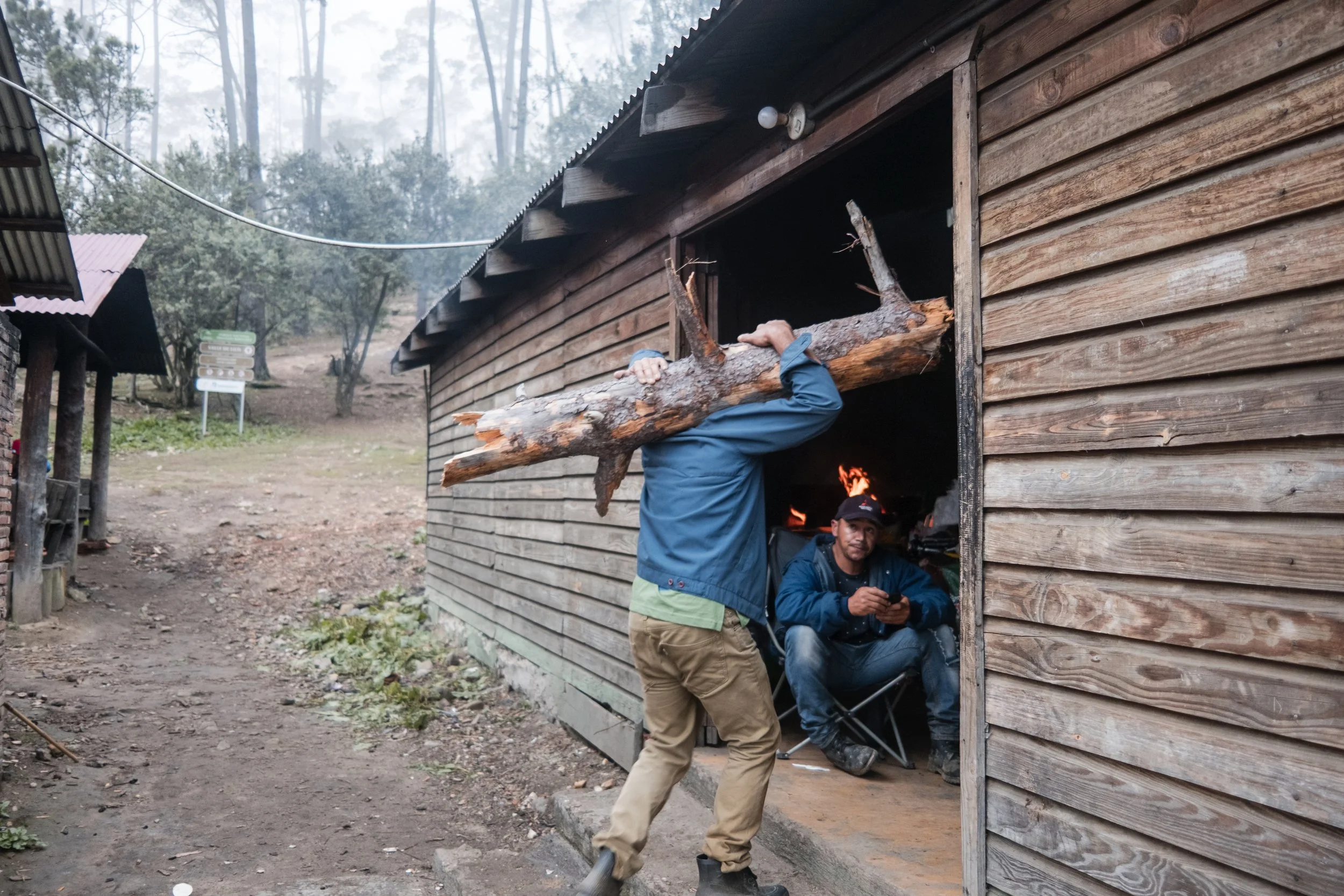 Hombre cargando un tronco de madera en la entrada de una cabaña, otra persona está sentada adentro cerca de un fuego. Bosque de fondo.