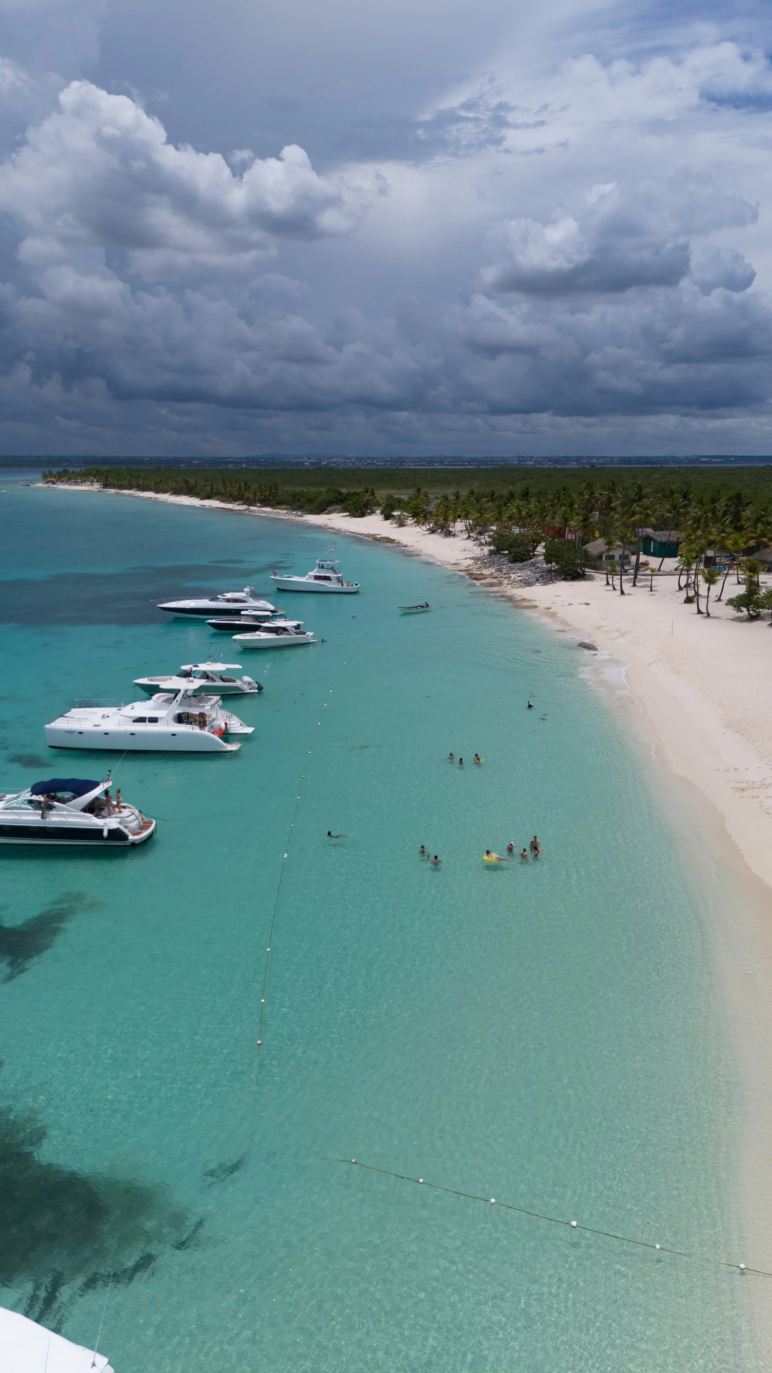 Playa tropical con arena blanca, agua turquesa y varias embarcaciones ancladas cerca de la orilla. Personas nadando en el agua. Cielo nublado en el fondo.