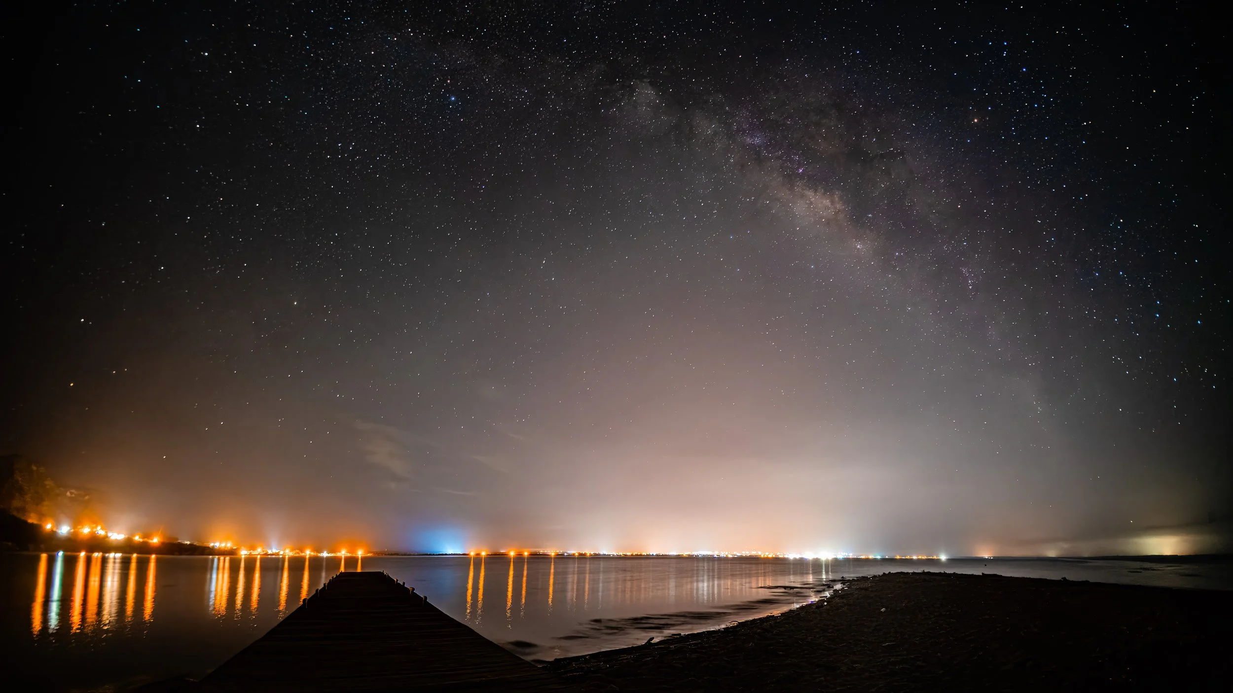 Cielo estrellado con la Vía Láctea sobre un muelle y luces de ciudad reflejándose en el agua.