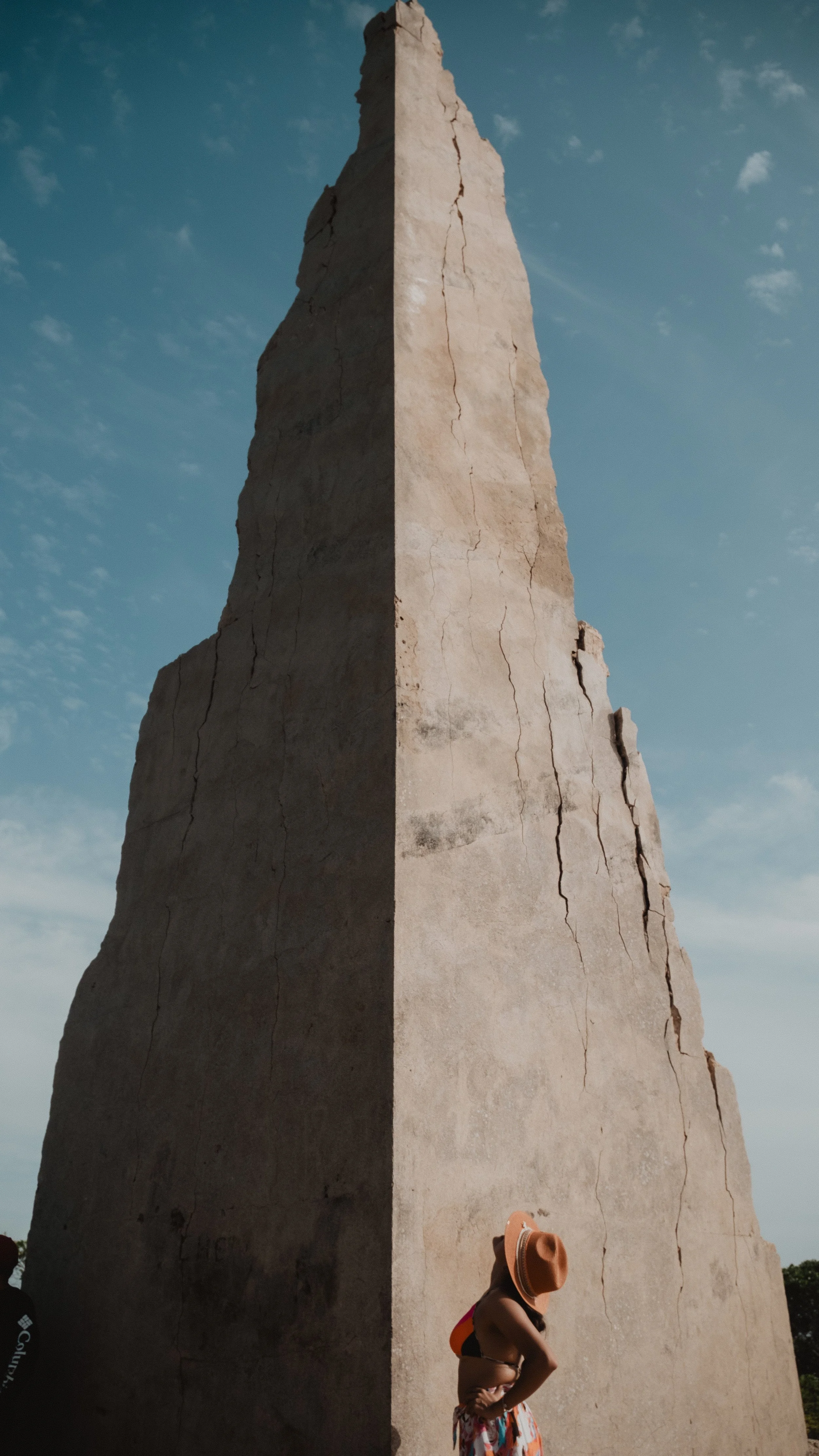 Una persona con sombrero junto a una gran estructura de piedra con forma puntiaguda y cielo azul de fondo.