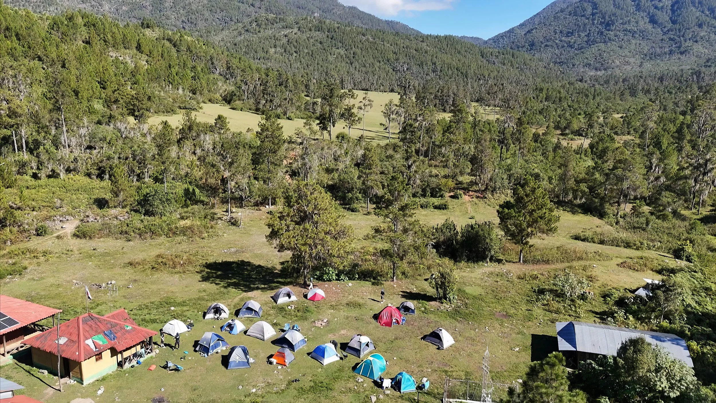Campamento con varias tiendas de campaña en un claro rodeado de bosque, con montañas al fondo.
