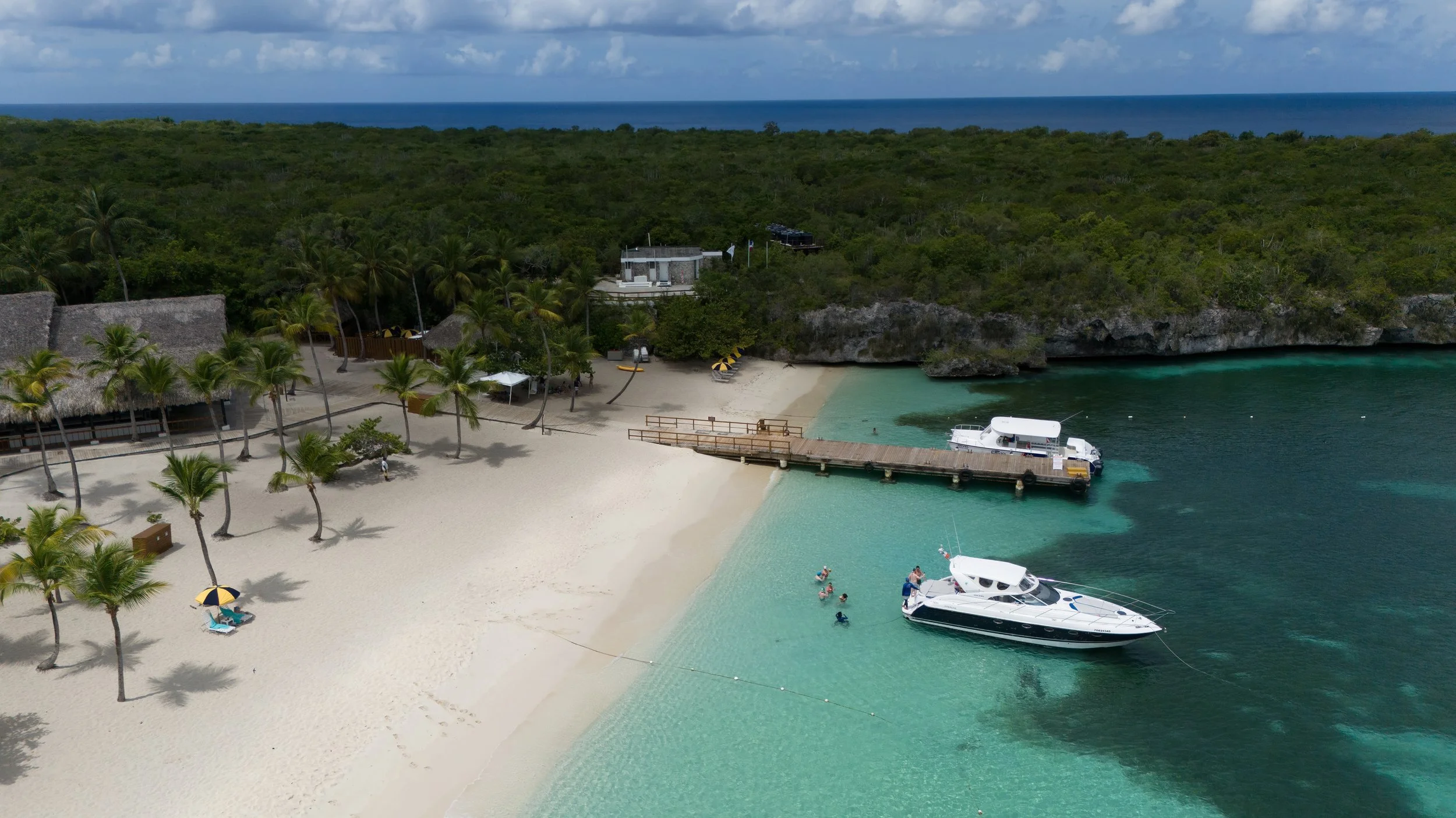 Vista aérea de una playa tropical con arena blanca y palmeras, un muelle de madera, un yate en el agua y personas nadando. Al fondo, un área boscosa y el mar.