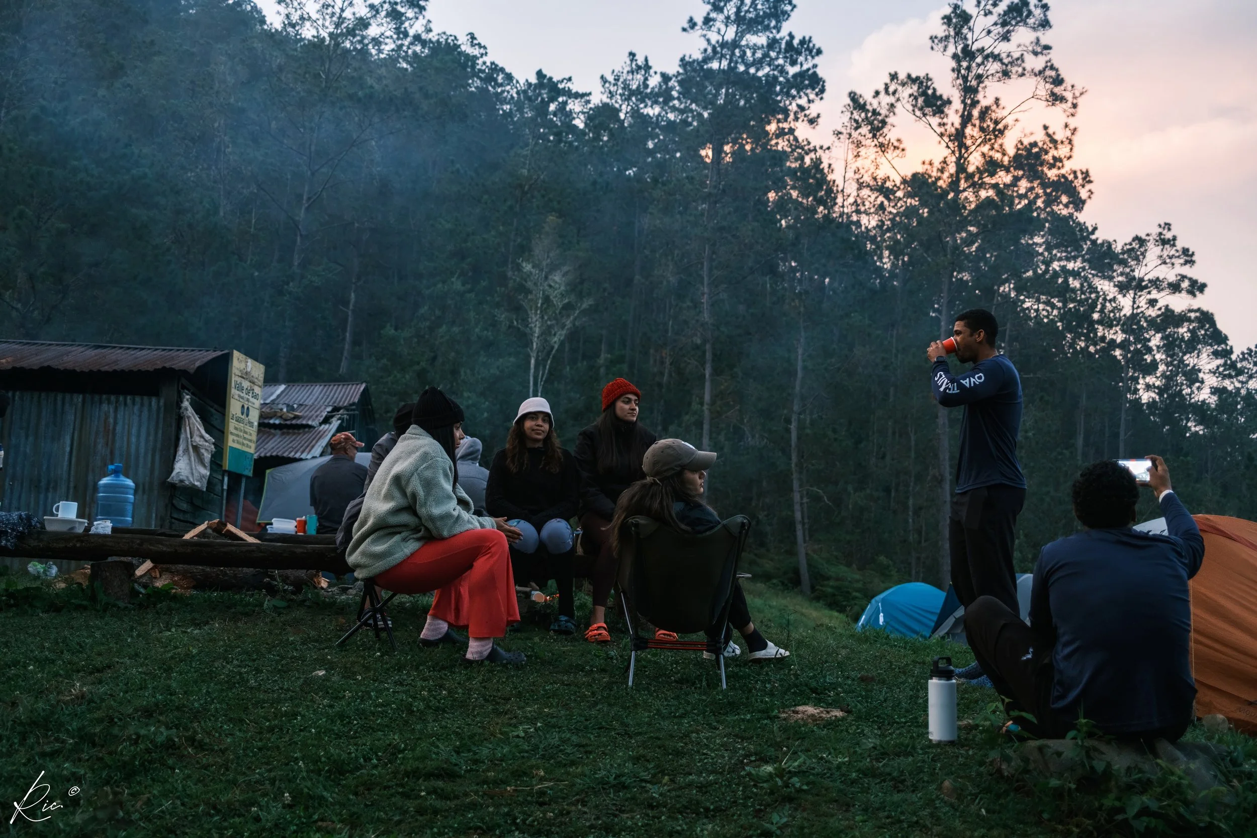 Grupo de personas en un campamento al aire libre durante el atardecer, rodeados de tiendas de campaña y un bosque de pinos, con una cabaña al fondo.