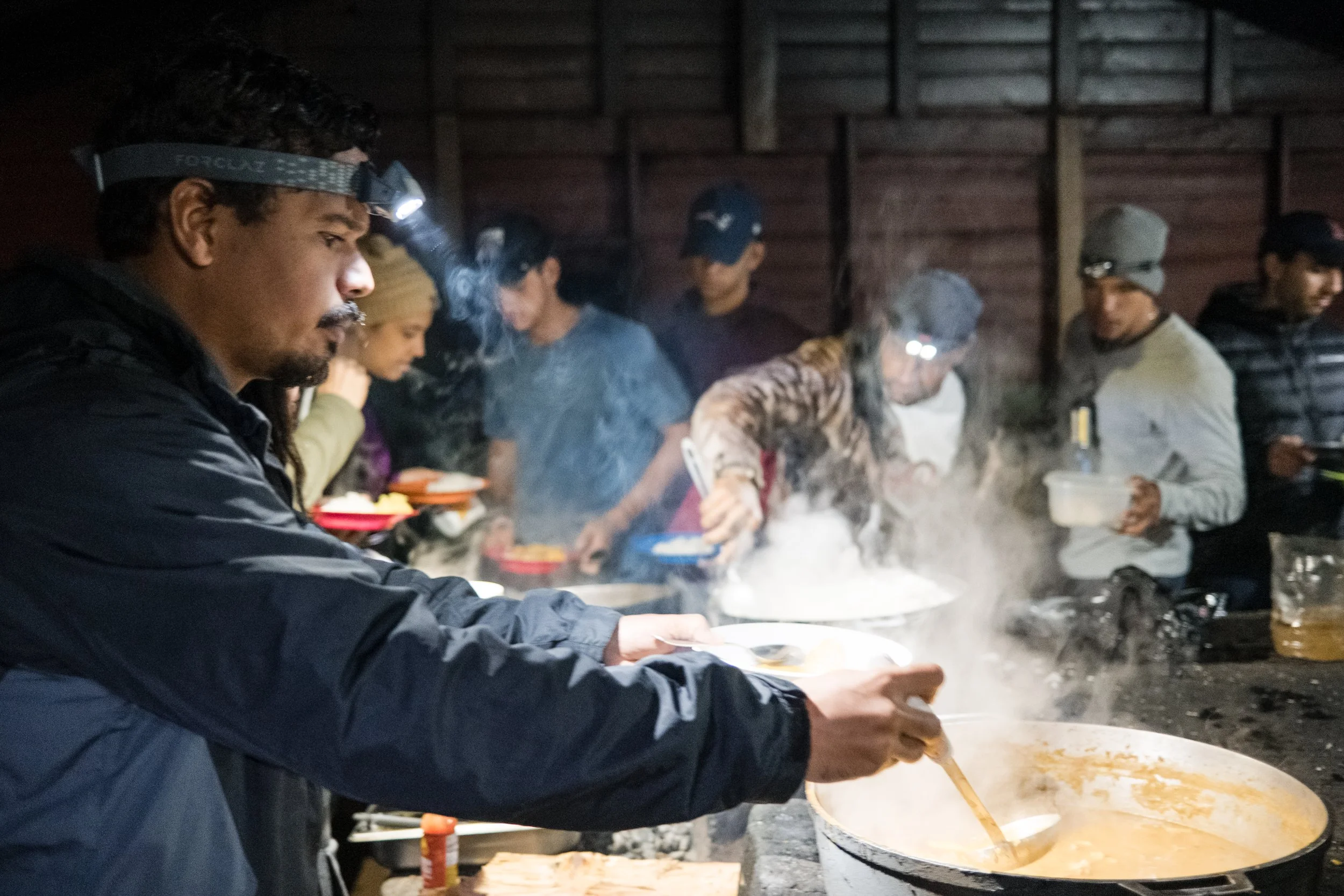Grupo de personas cocinando y sirviendo comida en un ambiente exterior, con linternas en la cabeza. Se observa una olla grande con sopa humeante.