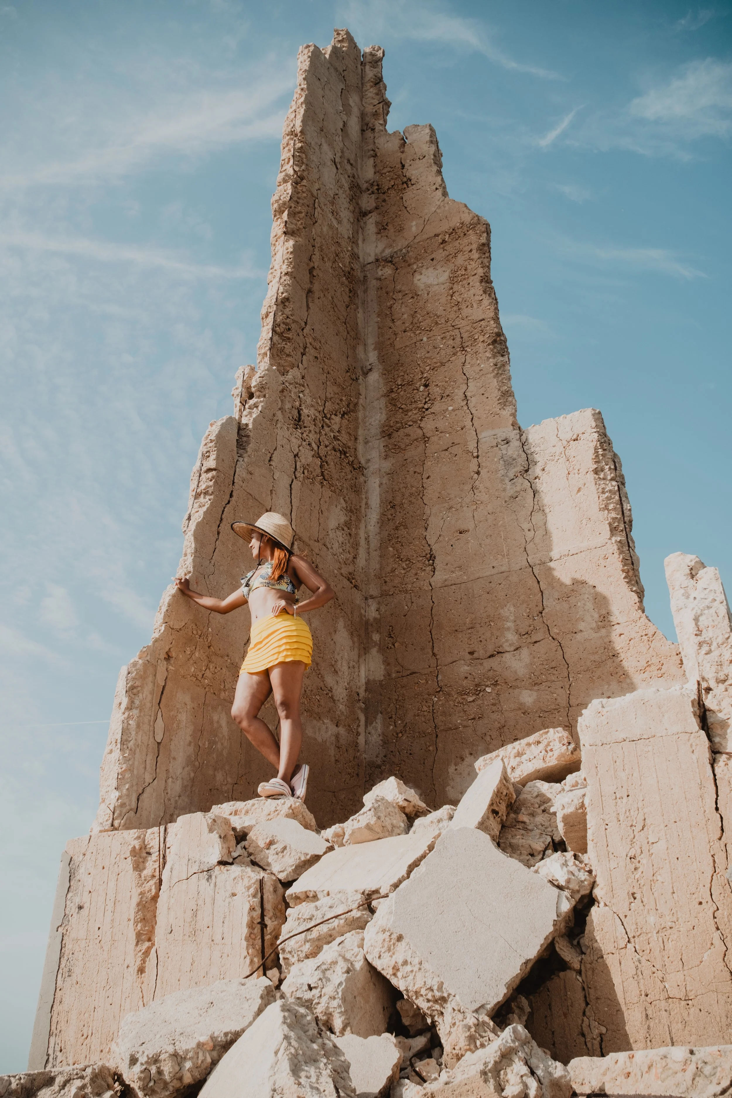 Una mujer con sombrero y falda amarilla sobre una estructura de ruinas bajo un cielo azul.
