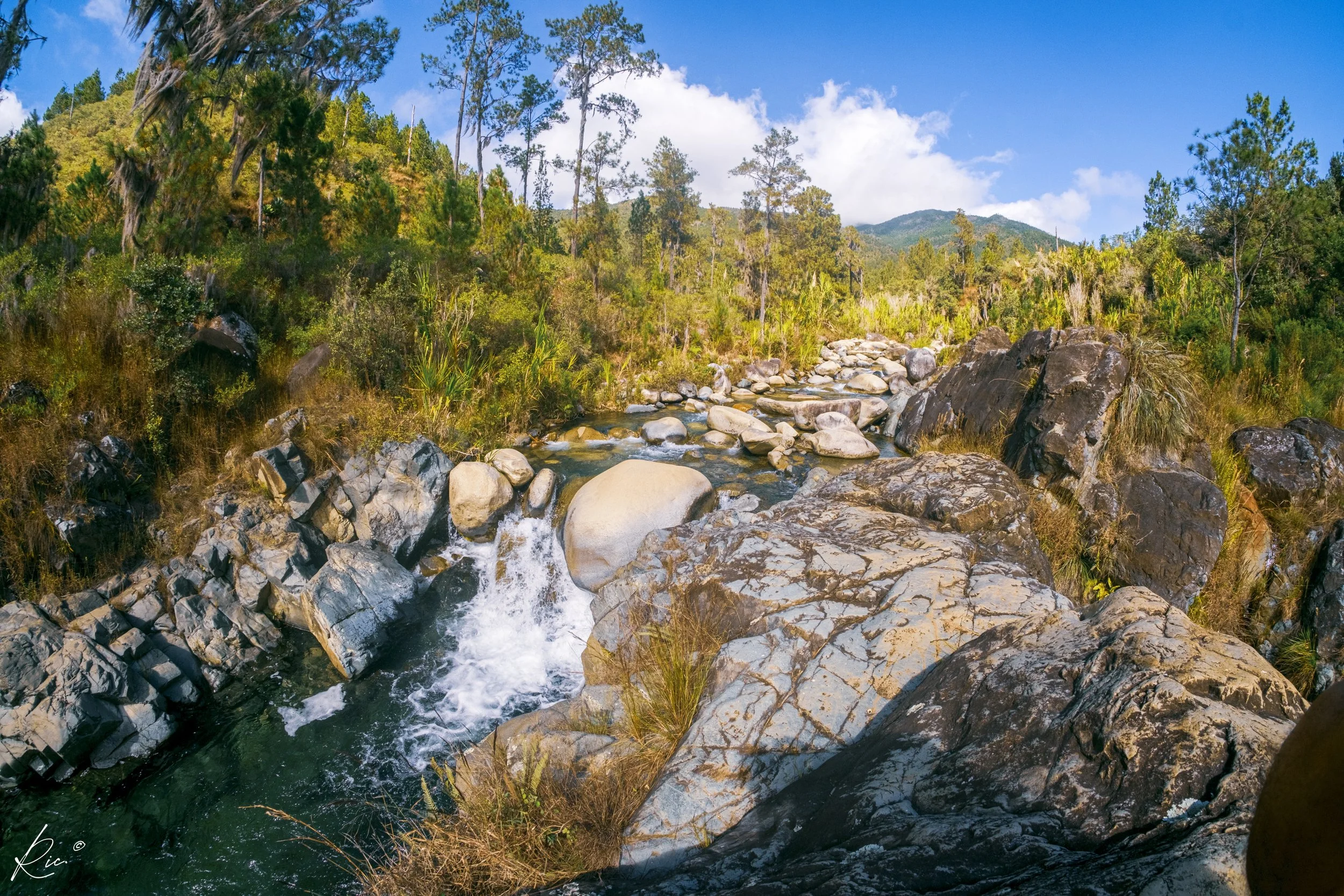 Paisaje natural con un río de aguas transparentes corriendo entre rocas grandes. Alrededor hay vegetación abundante y árboles sobre colinas. El cielo está despejado con algunas nubes. Ambiente de montaña con fresco y aire libre.