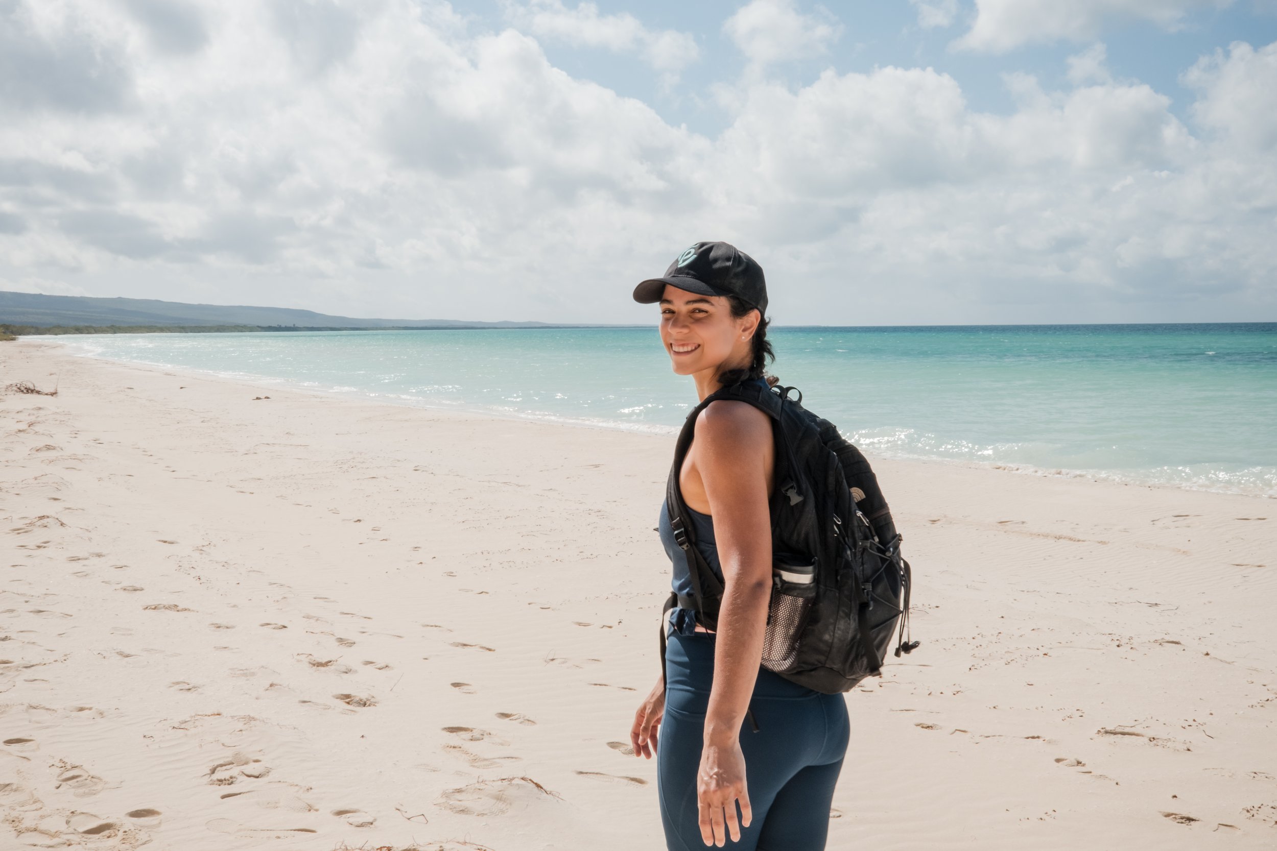 Mujer sonriente en la playa con mochila, mar azul y cielo nublado.