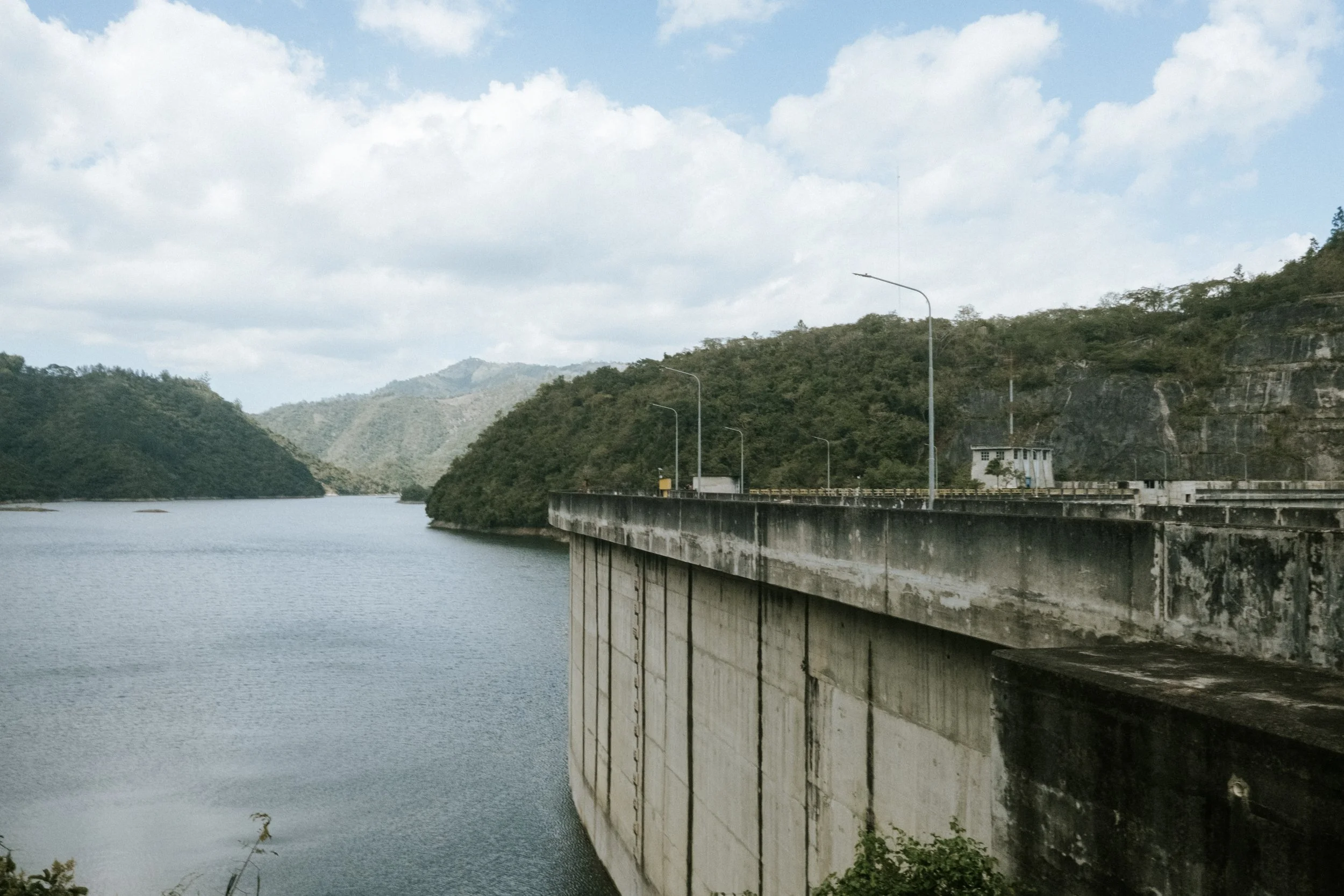 Presa de concreto con agua y montañas en el fondo bajo un cielo nublado.