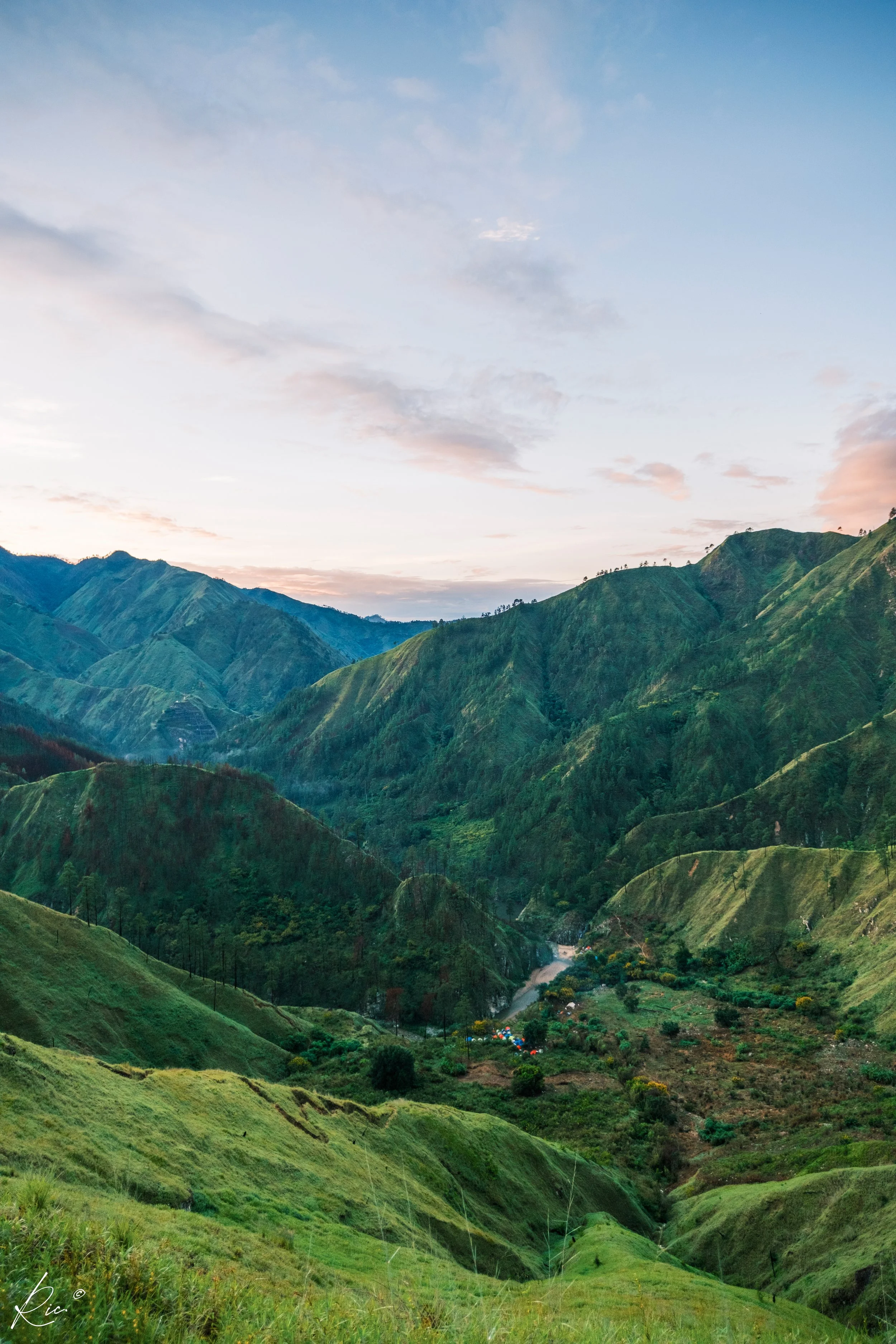 Paisaje montañoso verde con un valle al fondo y cielo despejado al amanecer.