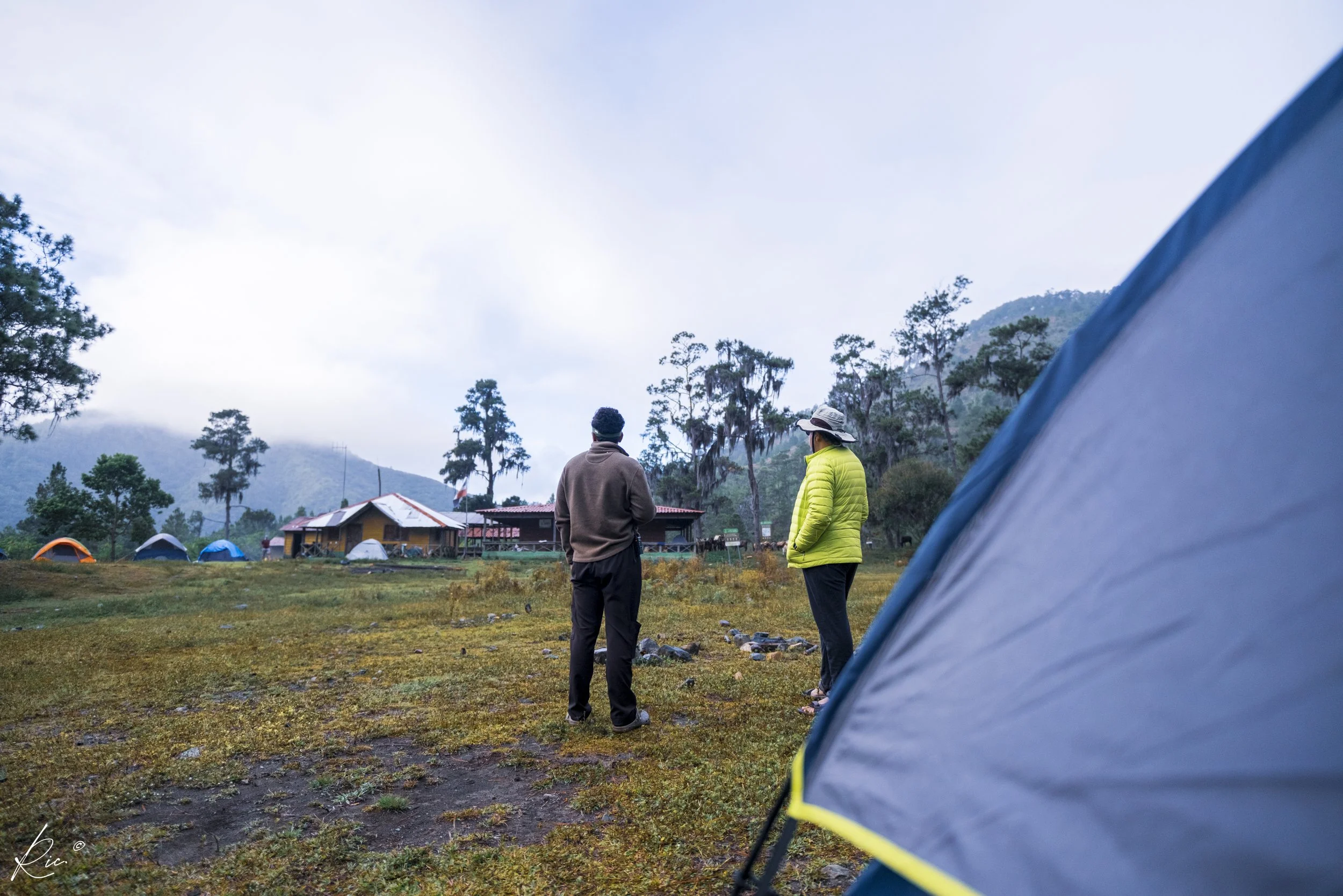 Personas al aire libre en un campamento, con una tienda de campaña en primer plano y un paisaje montañoso con árboles al fondo.