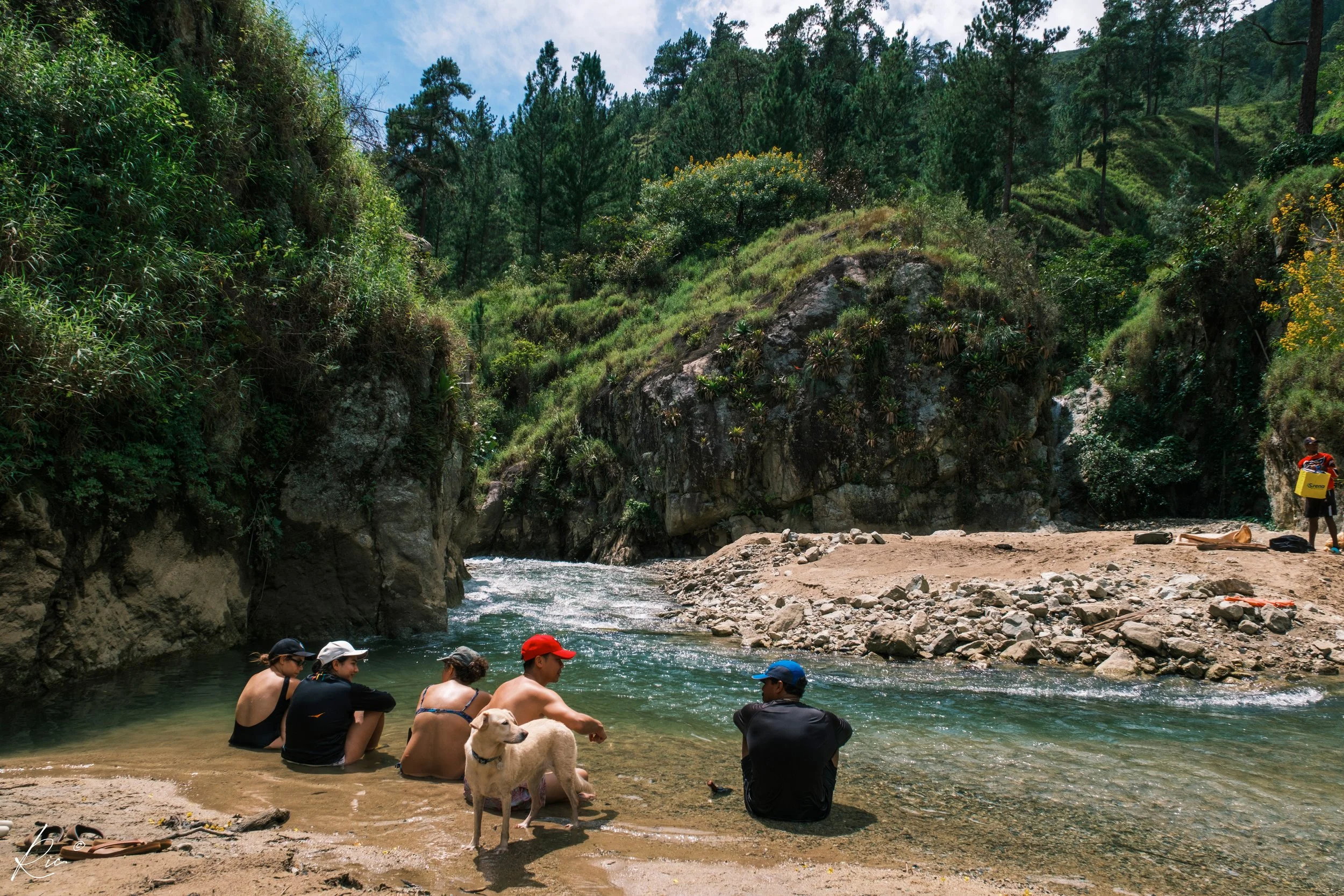 Personas sentadas junto a un río rodeado de montañas y vegetación, con un perro en la orilla.