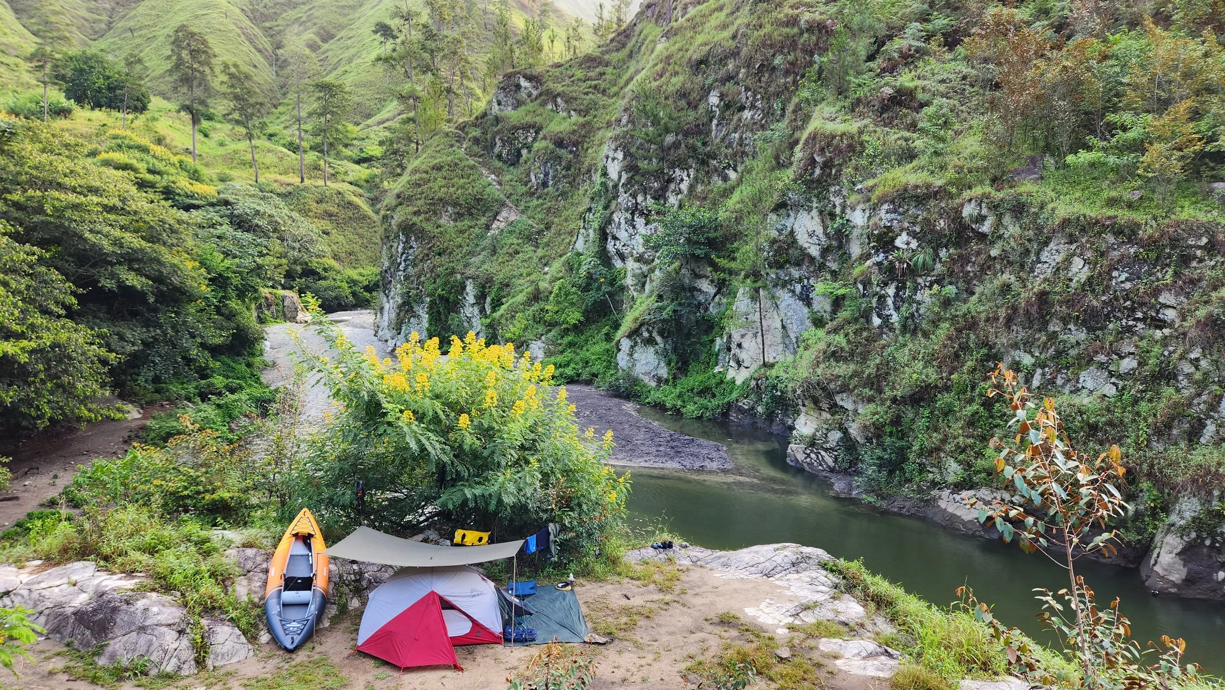 Campamento junto a un río en un valle montañoso, con tiendas de campaña, un kayak naranja y vegetación abundante.