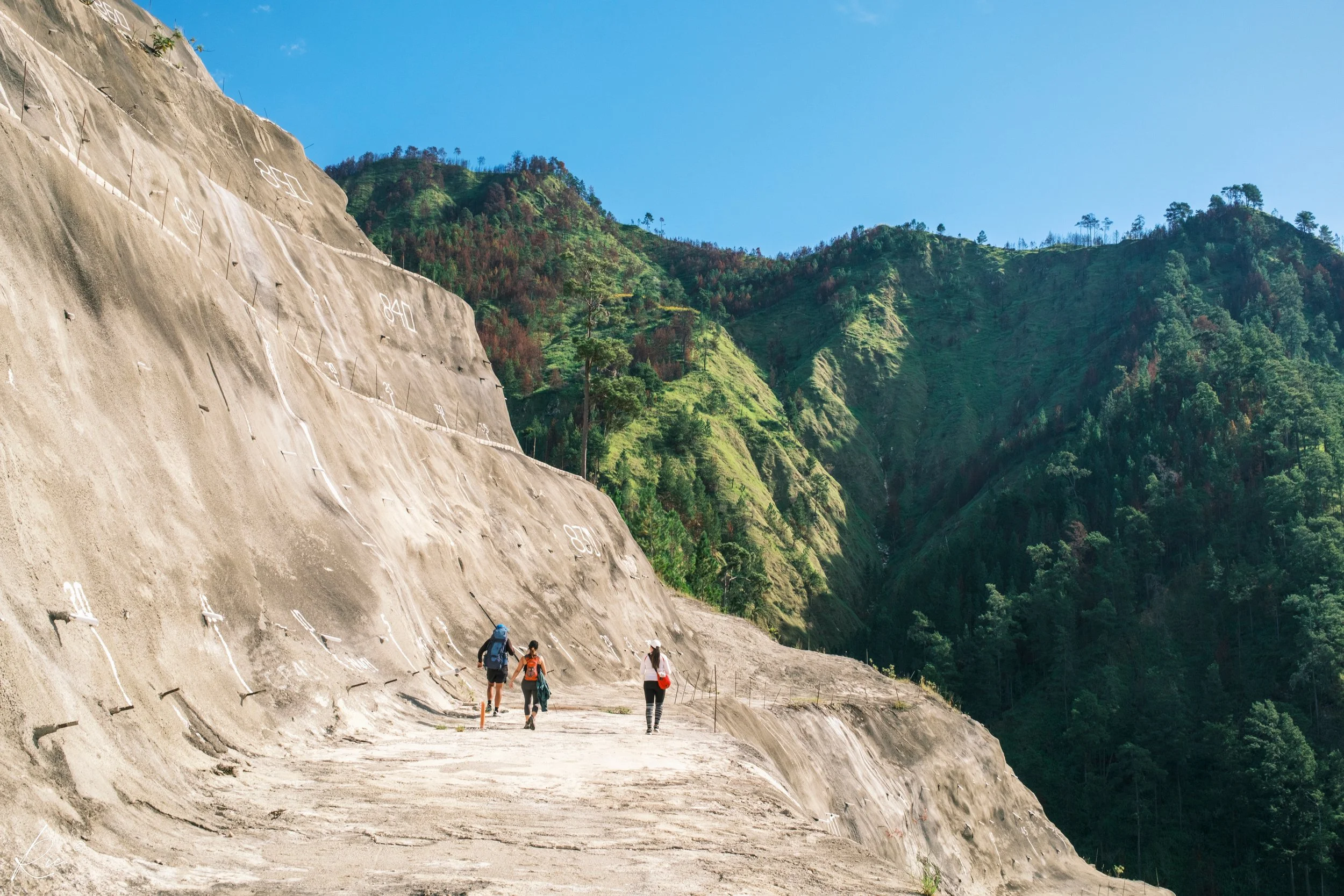 Tres personas caminando por un camino inclinado en una montaña, con vegetación verde en el fondo y un cielo despejado.