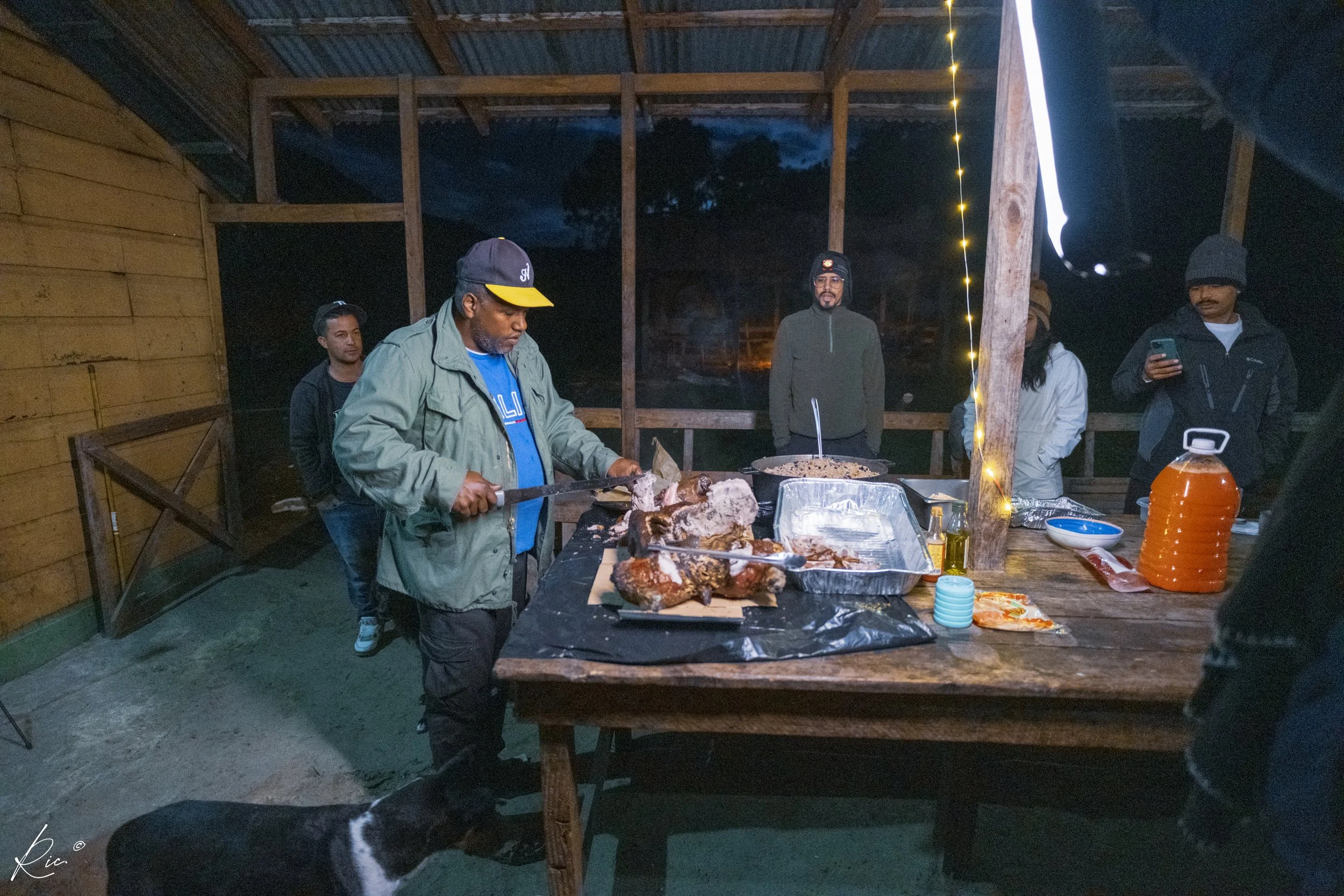 Grupo de personas en una barbacoa nocturna, una persona cortando carne en una mesa con luces decorativas alrededor.