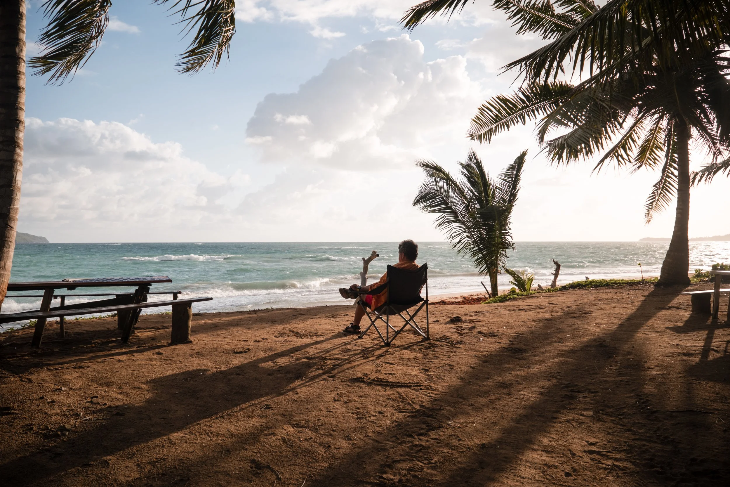 Persona sentada en una silla de playa bajo palmeras, mirando el mar en una playa al atardecer.