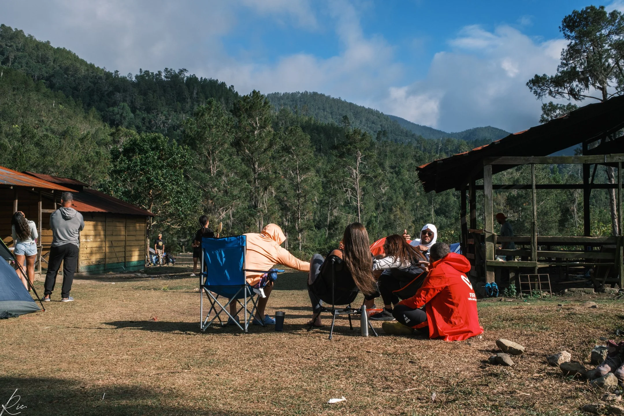 Personas sentadas y de pie en un campamento al aire libre, rodeadas de montañas y árboles, con una cabaña de madera y una estructura de techo de teja cerca.