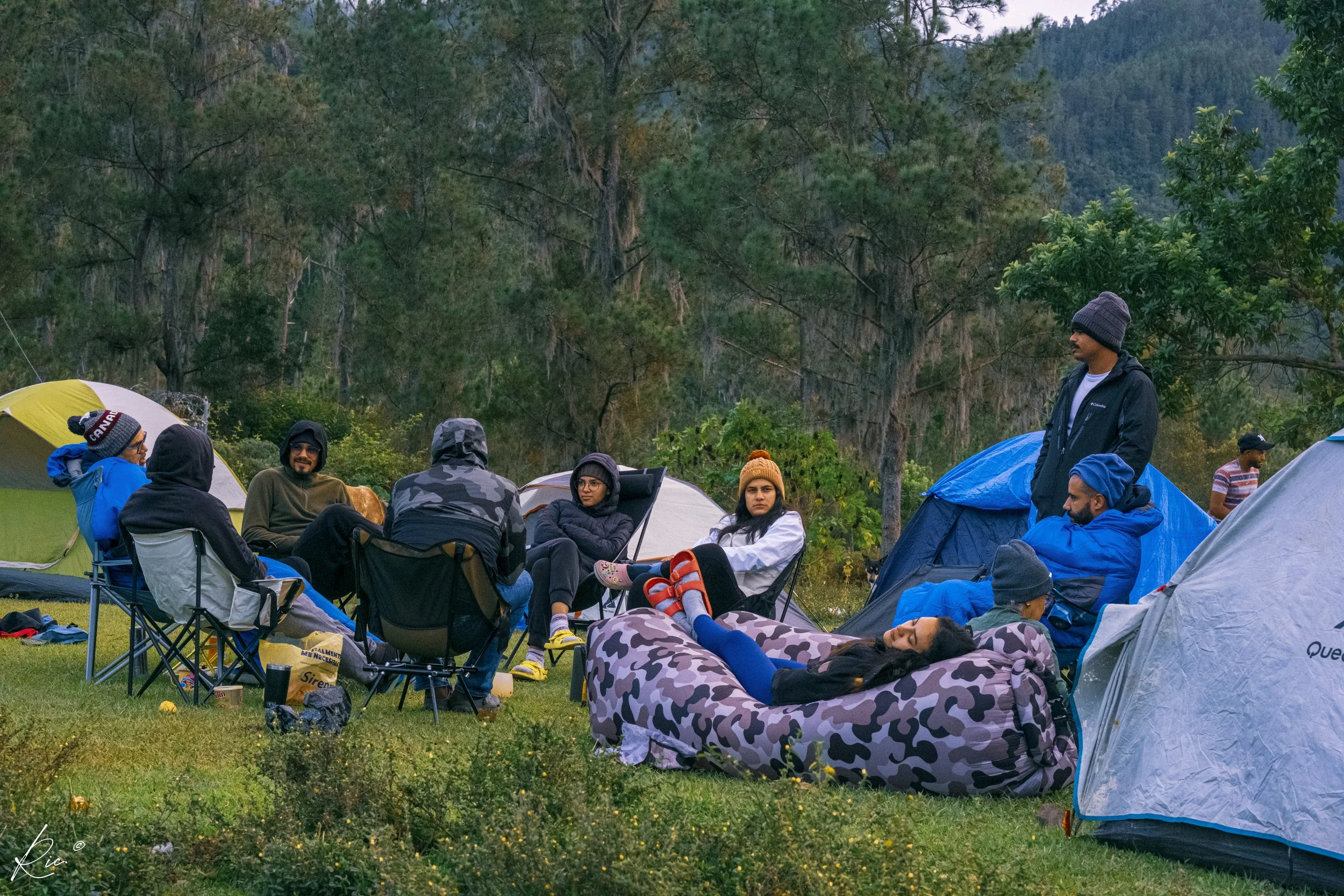 Grupo de personas sentadas alrededor de una fogata en un campamento con tiendas de campaña en un bosque.