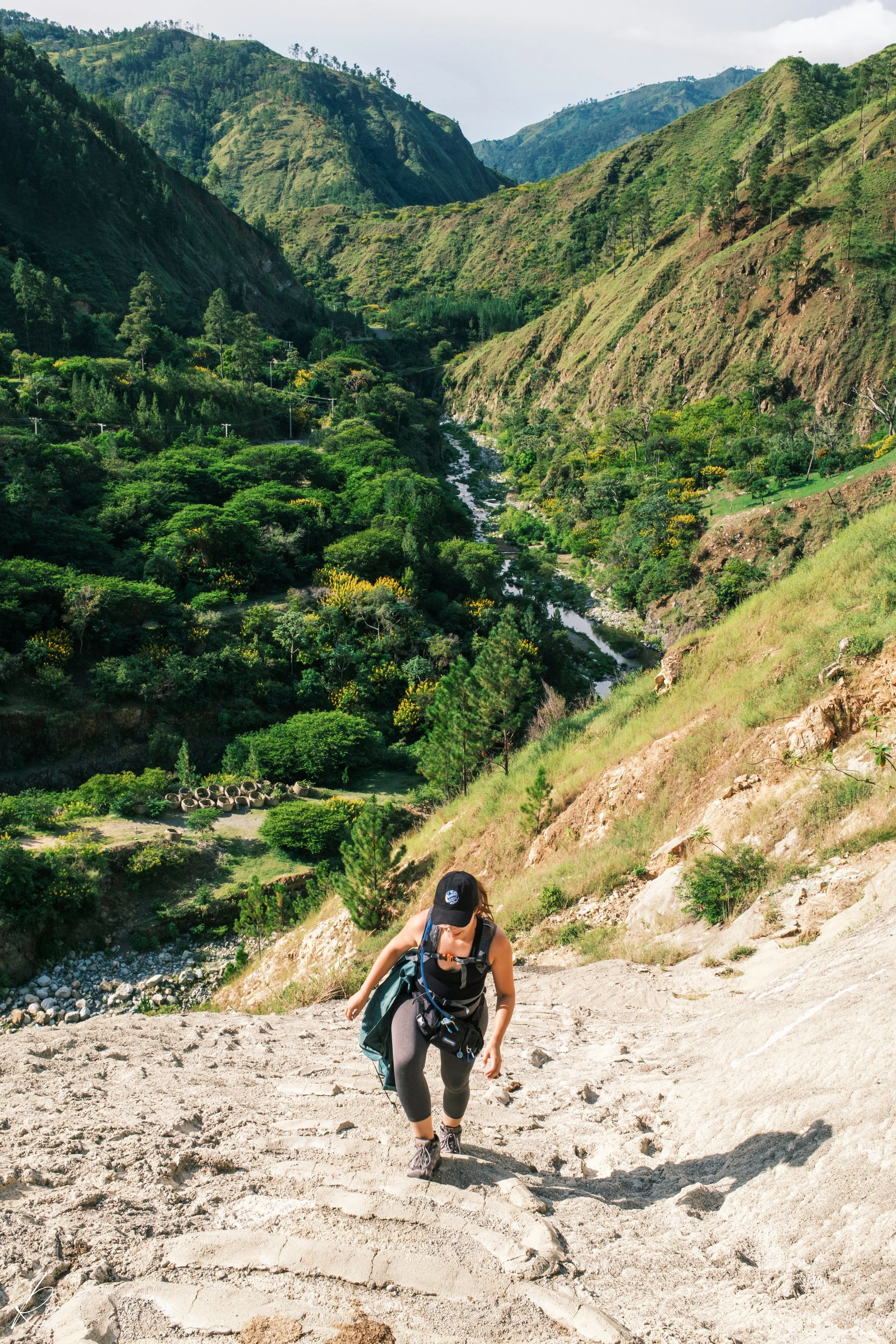 Persona subiendo una colina arenosa con un paisaje montañoso y un río al fondo.