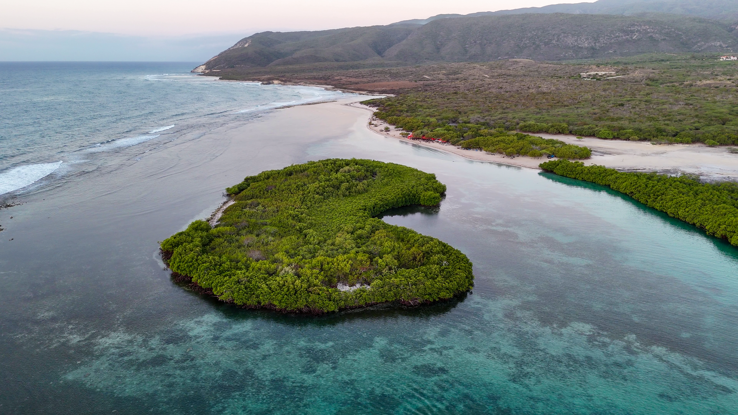 Paisaje costero con una isla cubierta de manglares, agua turquesa y montañas en el fondo.