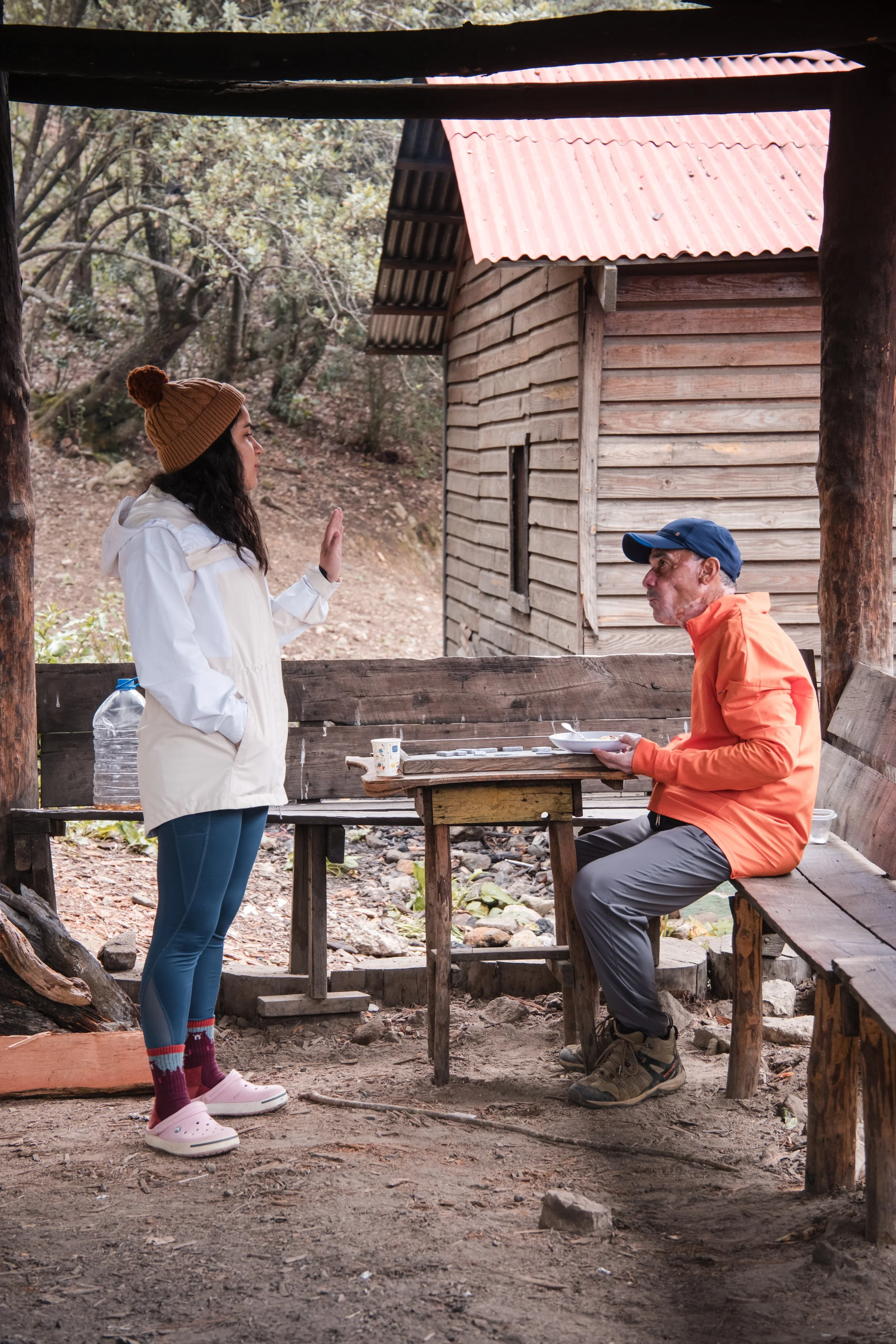 Dos personas en un refugio de madera en el bosque, una está sentada en un banco comiendo, la otra está de pie gesticulando. Hay una mesa improvisada con algunos objetos sobre ella.