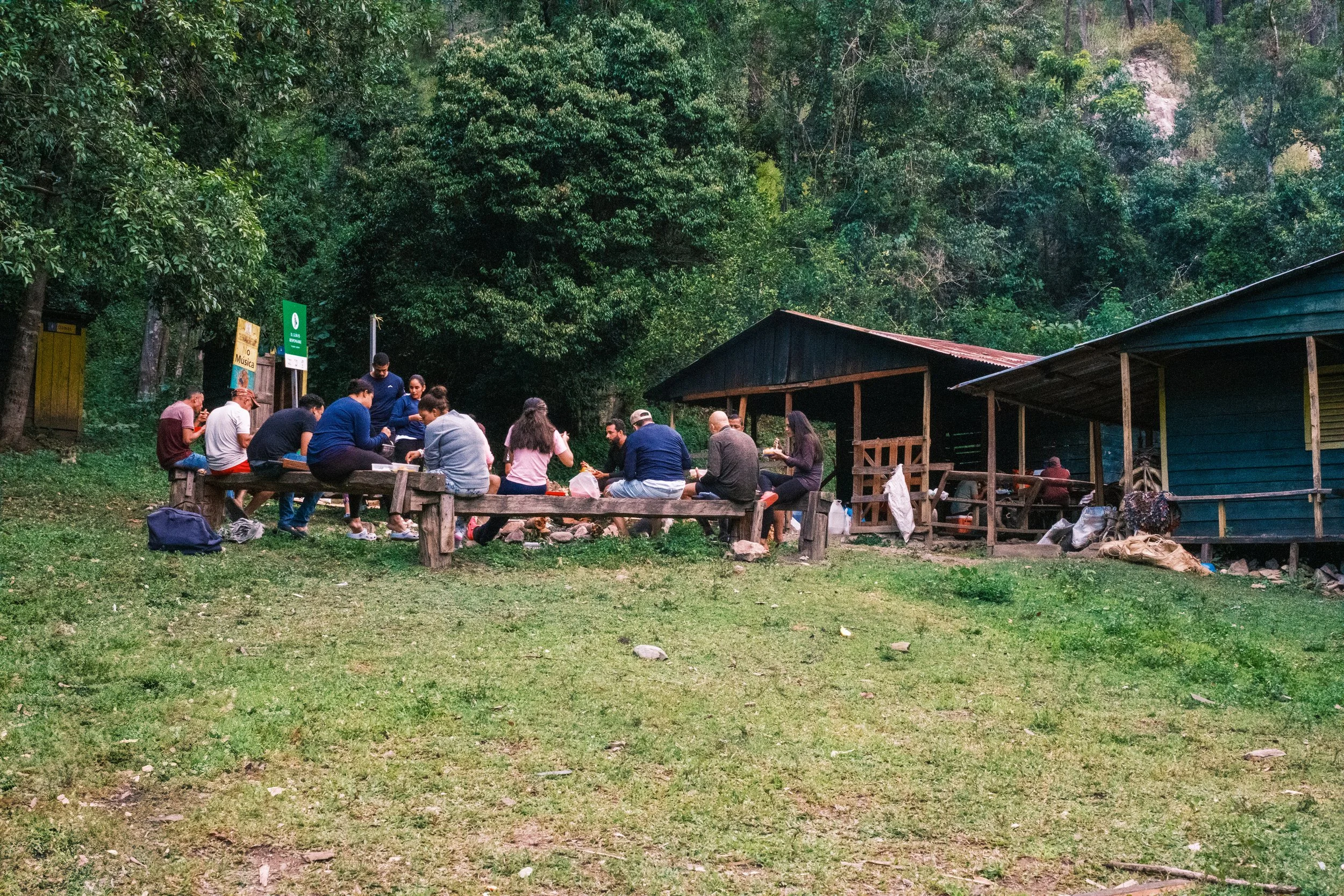 Grupo de personas sentadas en bancos al aire libre, rodeadas de árboles y cabañas de madera.
