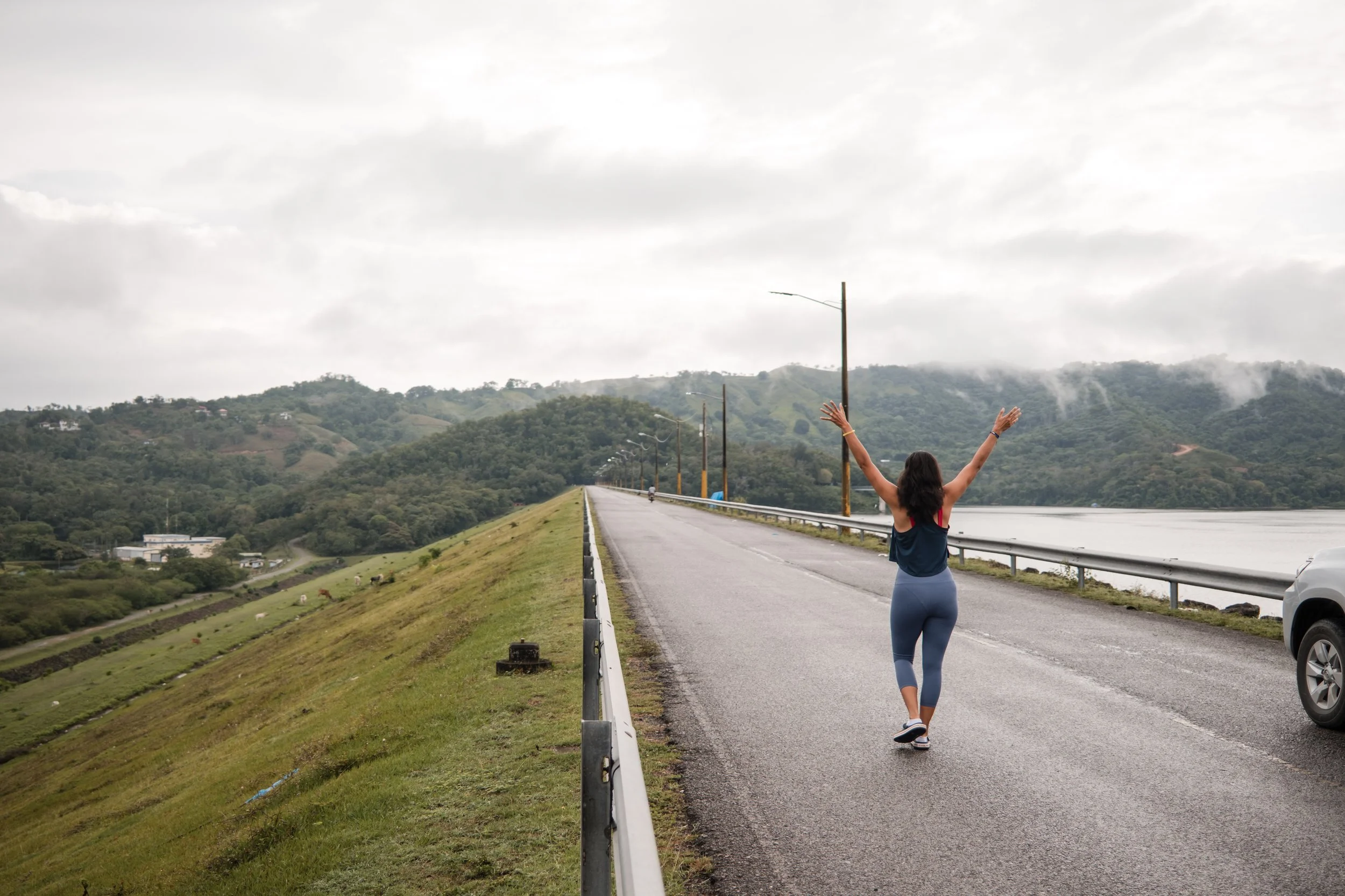 Mujer caminando con los brazos levantados por una carretera al lado de un lago y montañas con nubes en el fondo.